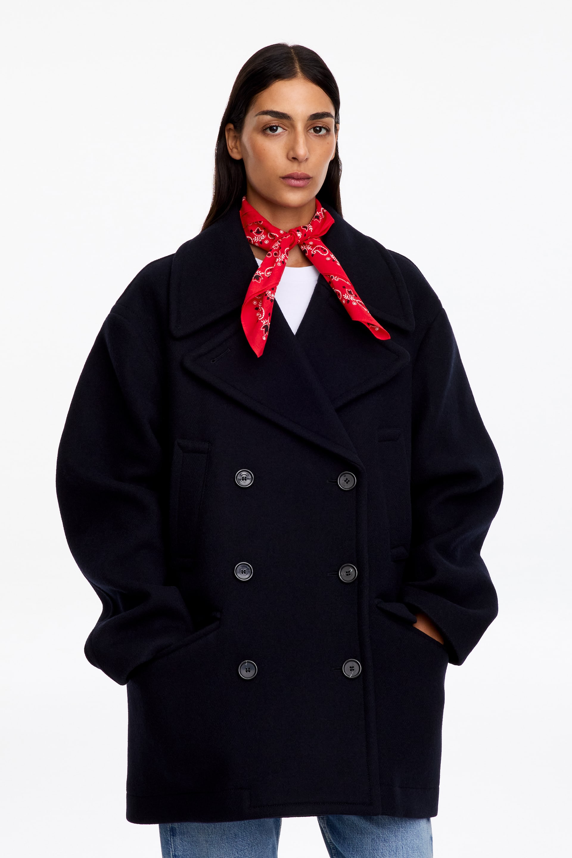 Vezi imaginea mai mare: O eșarfă roșie din bumbac, cu un model paisley alb și negru, este purtată legată într-un nod în jurul gâtului modelului. Eșarfa este stilizată peste un tricou alb și purtată în straturi sub un palton bleumarin închis, cu închidere dublă la nasturi.