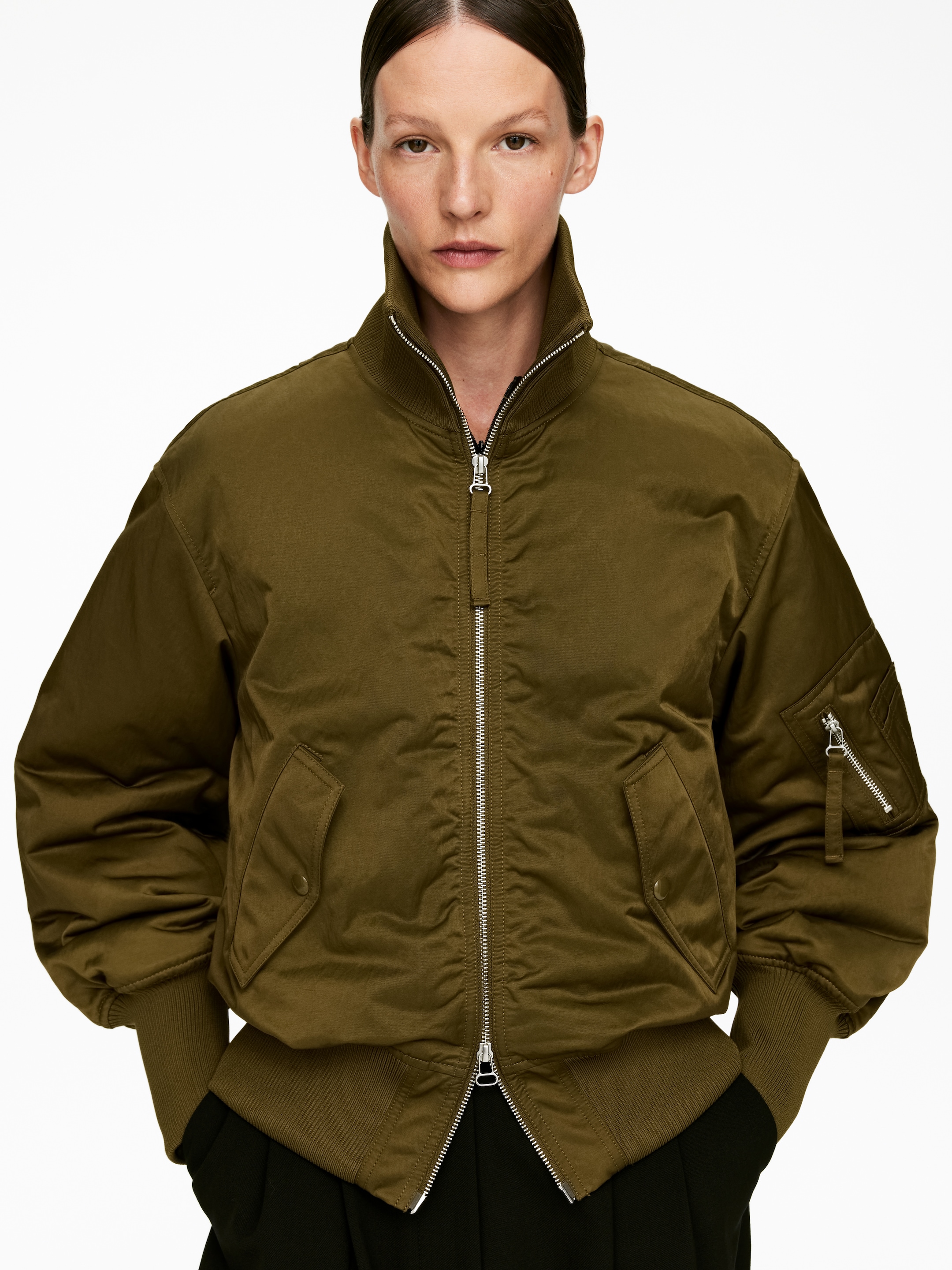 Grösseres Bild ansehen: Eine olivgrüne Bomberjacke wird von vorne gezeigt, mit einem gerippten Stehkragen, einem silbernen Reißverschluss in voller Länge und einer übergroßen Passform mit voluminösen Ärmeln. Sie präsentiert auch zwei Klappentaschen mit Druckknopfverschlüssen, gerippte Bündchen, einen gerippten Saum und eine Reißverschlusstasche auf dem linken Ärmel.