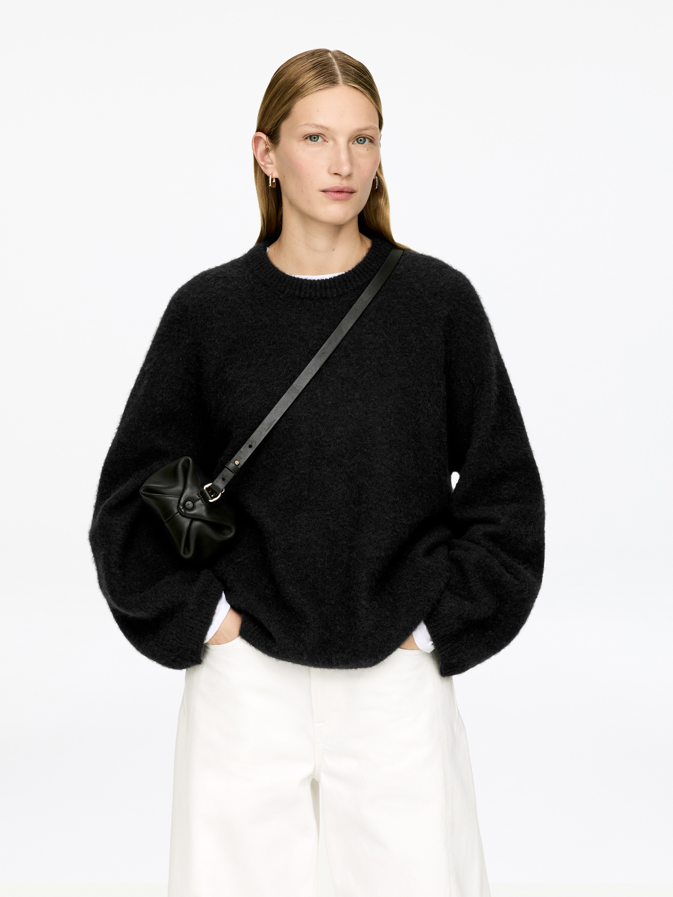 Grösseres Bild ansehen: Ein schwarzer, gebürsteter Strickpullover mit Rundhalsausschnitt wird getragen und von vorne gezeigt. Er hat eine lockere Passform mit voluminösen Ärmeln und breiten Bündchen, und ein weißes Hemd ist sichtbar, das am Ausschnitt und Saum darunter liegt.