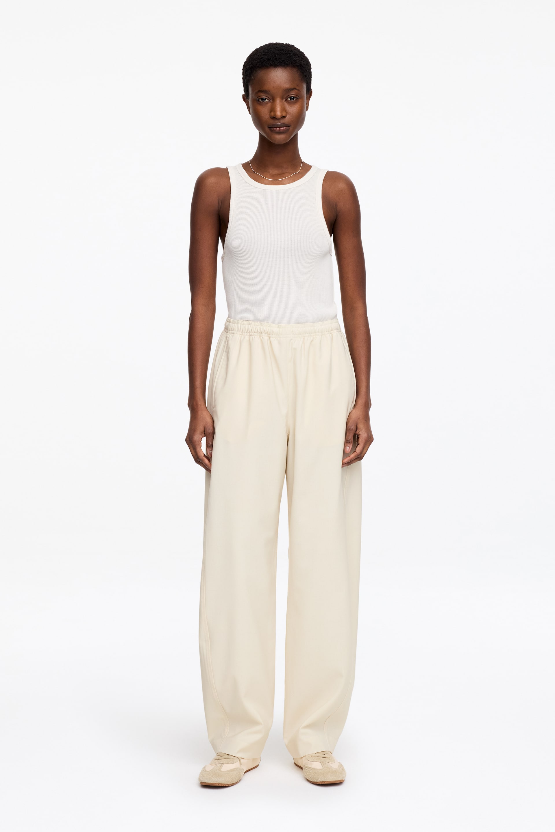 Ingrandisci l'immagine: Una canotta bianca a coste, caratterizzata da uno scollo rotondo e una silhouette aderente, è presentata infilata in pantaloni a gamba larga color crema, dotati di una vita elasticizzata e tasche laterali. L'insieme è rivolto in avanti.