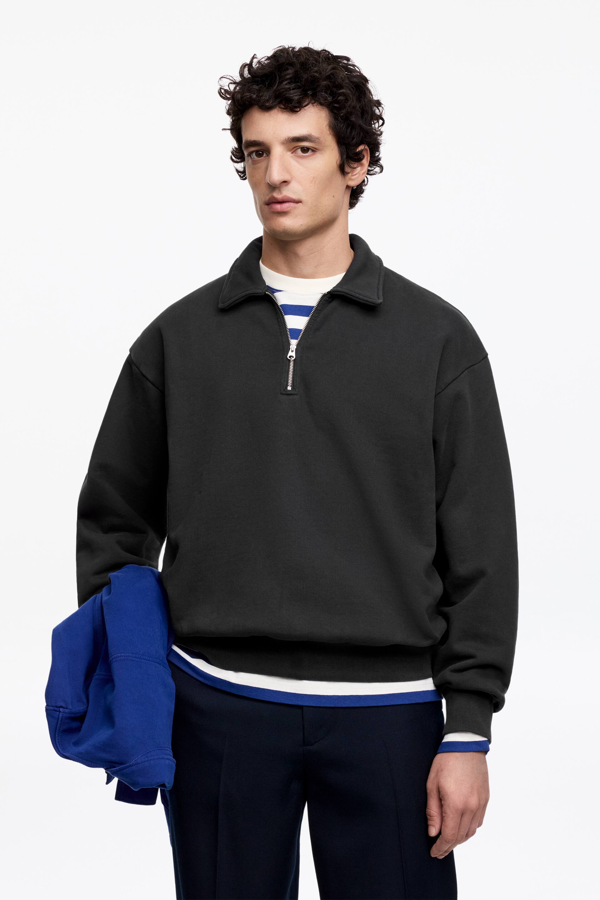 Näytä suurempi kuva: Musta neljännesvetoketjullinen pusero esitellään, jossa on levitetty kaulus, laskettu olkapäät, ribatut hihansuut ja ribattu helma. Valkoinen ja siniraidallinen paita näkyy kauluksen, hihansuiden ja helman alta.