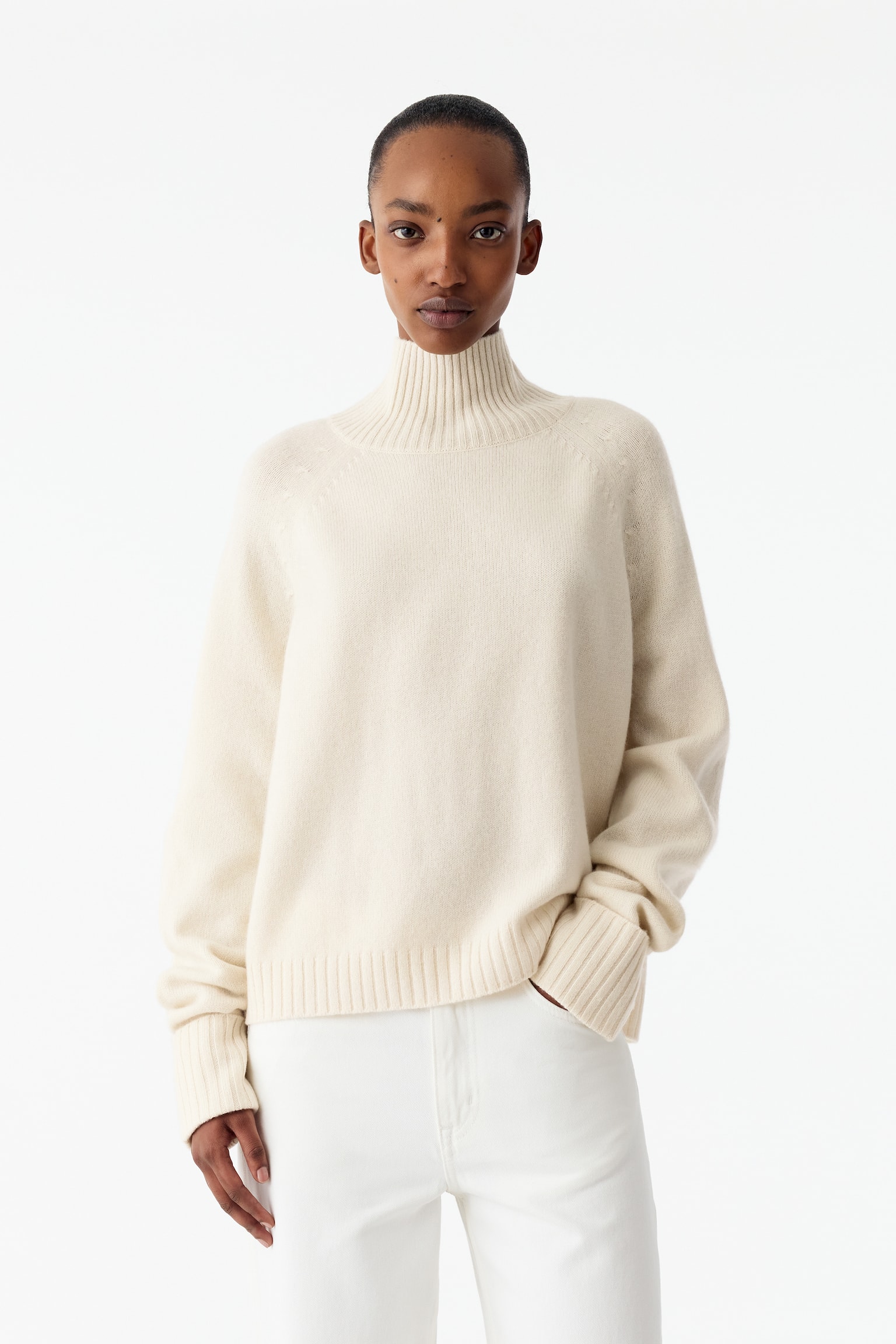 En off-white strikket høyhalset genser bæres av modellen, med raglanermer, nedbrettede ribbestrikkede mansjetter og ribbestrikket kant. Genseren er stylet med hvite bukser, og modellen har den ene hånden i en lomme. - 1