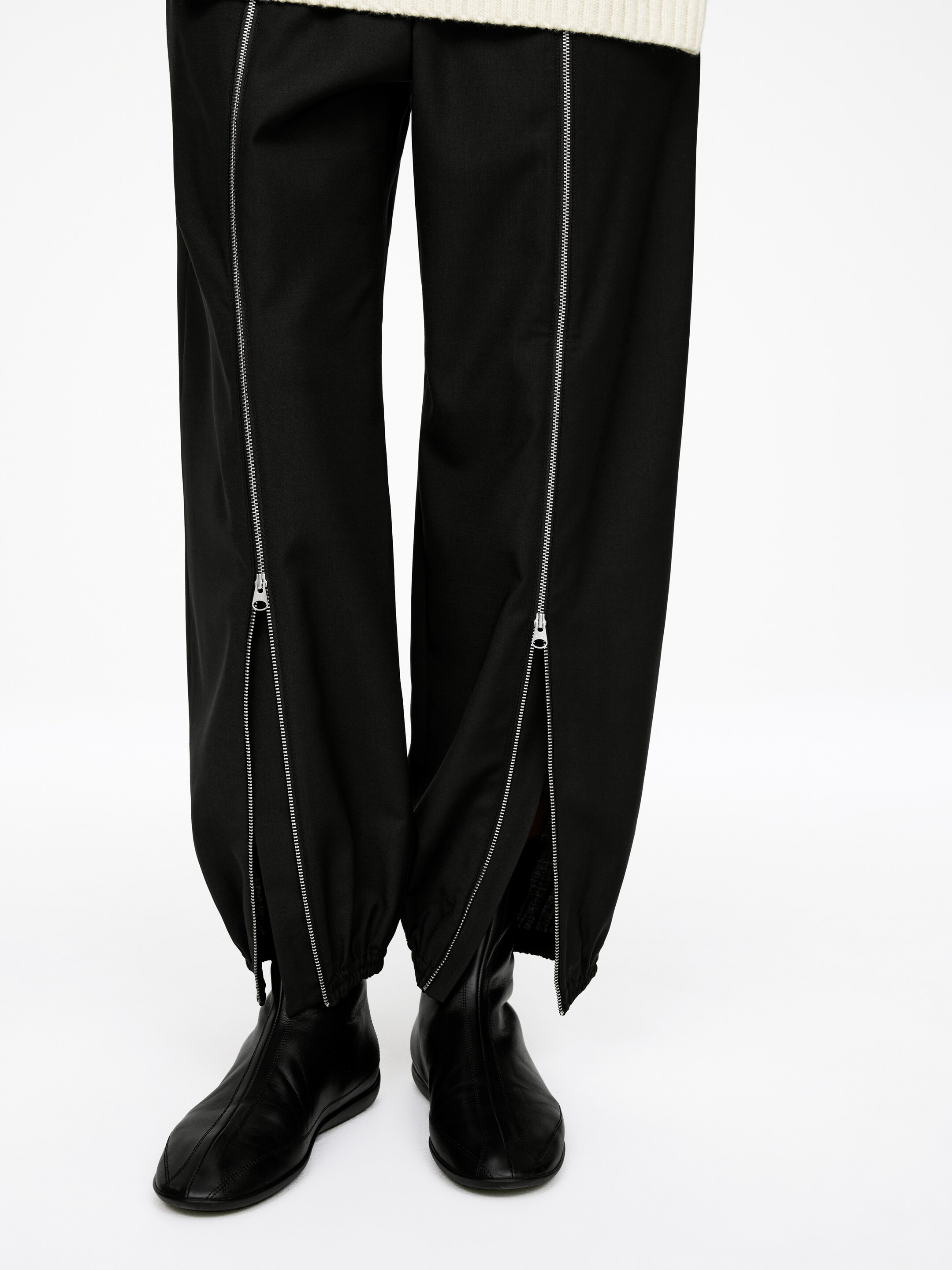 View larger image: Black pants are featuring full-length silver zippers running vertically down the front of each leg. The zippers at the hem are being unzipped, creating an open ankle detail and revealing black boots underneath.