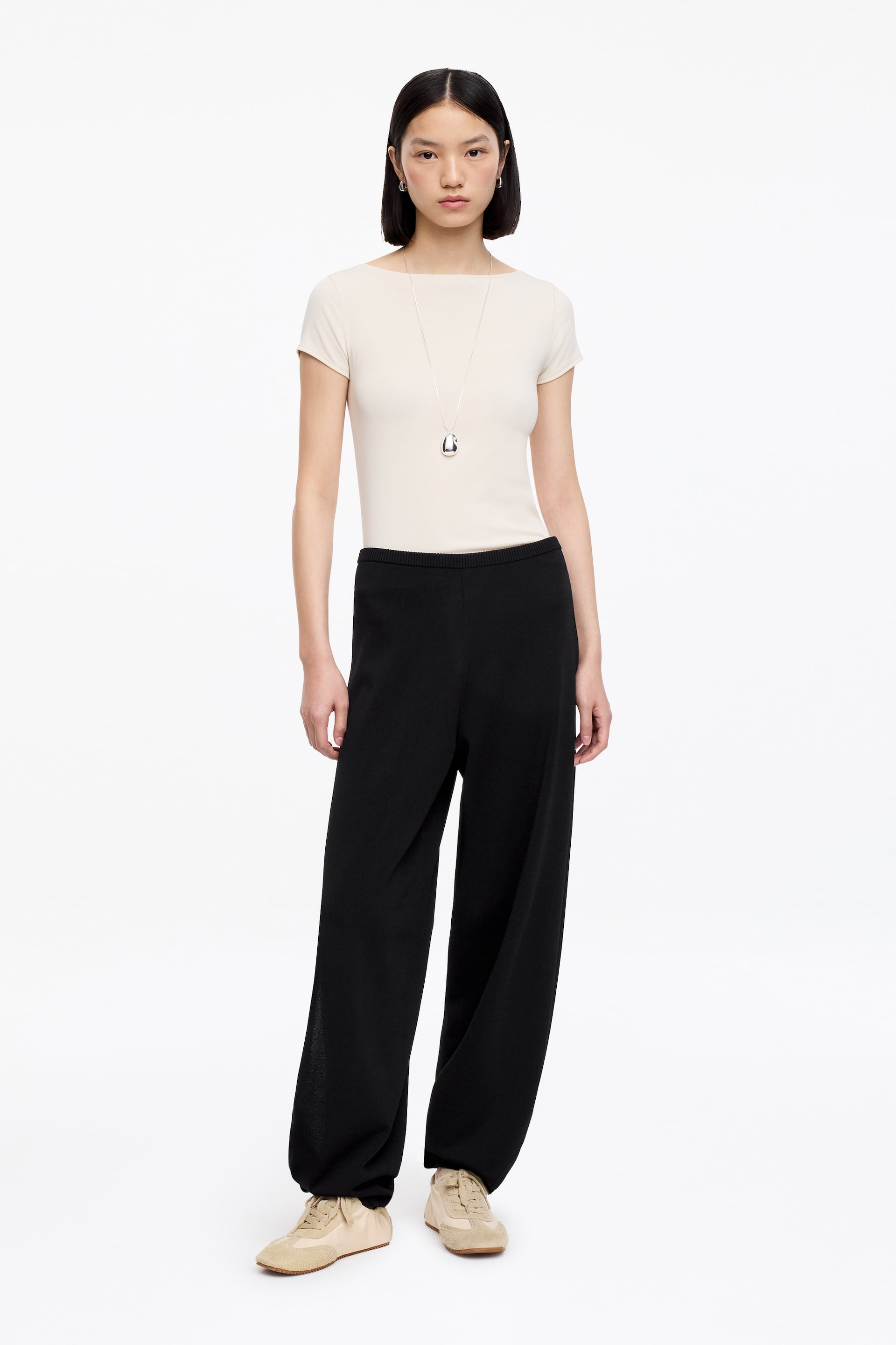 Visa större bild: Avslappnade stickade byxor i svart bärs av modellen, med en avslappnad, vid silhuett som smalnar av vid anklarna. Byxorna stylas med en krämfärgad kortärmad topp instoppad i midjebandet och beige sneakers.