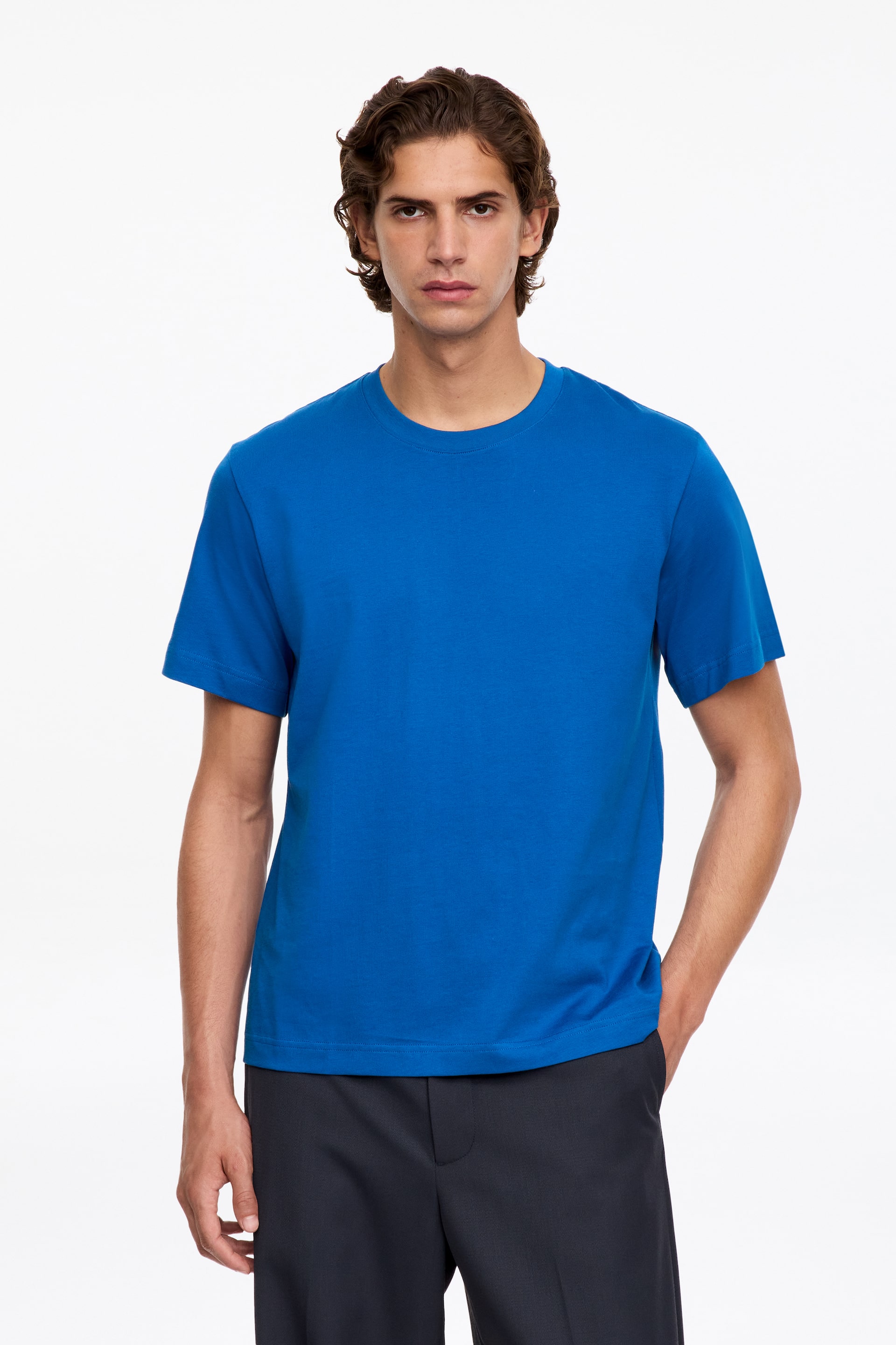 Vezi imaginea mai mare: Un tricou cu guler rotund, albastru aprins și cu mânecă scurtă, este purtat de model. Tricoul are o croială clasică, fluidă, și este asortat cu pantaloni gri închis.
