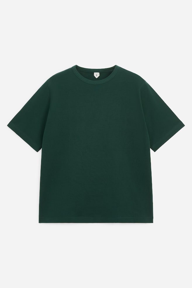 Ciemnozielony T-shirt z okrÄ
gÅym dekoltem jest pokazany z przodu, wyróżniajÄ
c siÄ swobodnym, lekko oversizeâowym krojem z szerokimi, wydÅużonymi krótkimi rÄkawami. Koszulka zostaÅa wykoÅczona klasycznym prÄ
żkowanym ÅciÄ
gaczem przy dekolcie oraz prostym doÅem. - 1