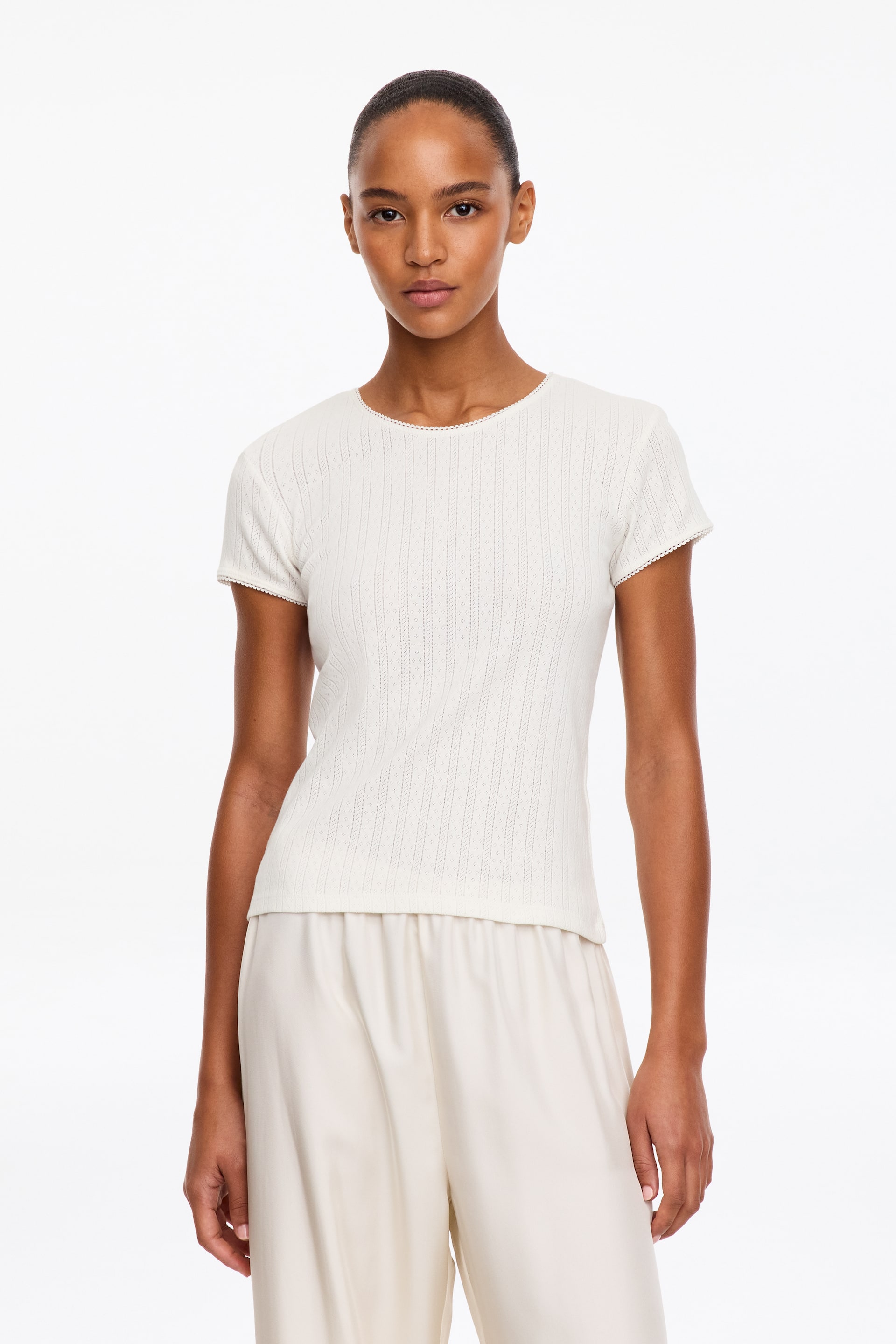 Ver imagen más grande: La modelo lleva un top de punto pointelle de color crema, de manga corta, con cuello redondo y delicado acabado de picot. El top tiene un ajuste regular y se combina con un pantalón fluido de color claro.