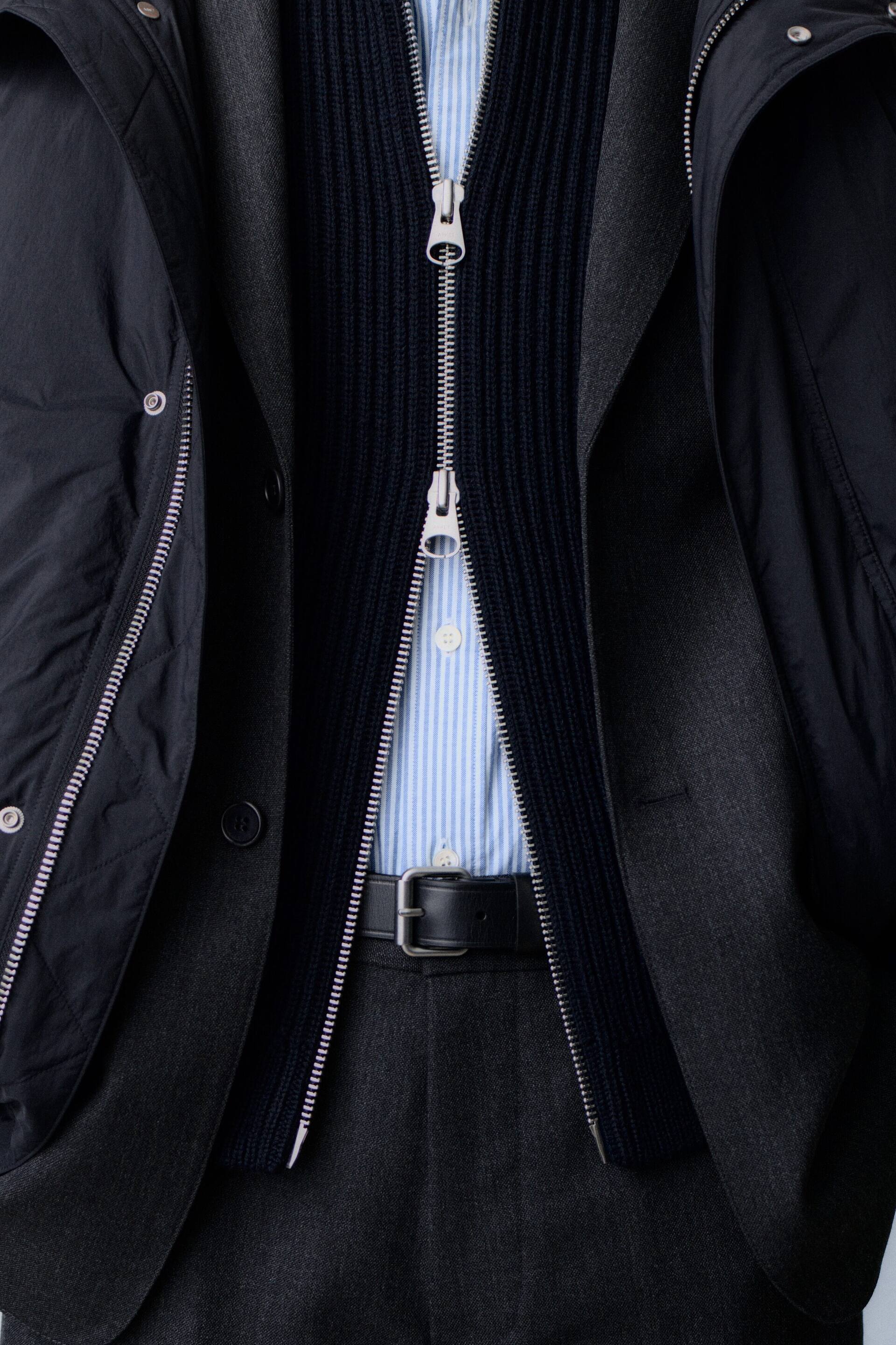 Visualizza immagine più grande: Una vista frontale mette in risalto un outfit multistrato, composto da una camicia a righe azzurre e bianche infilata in pantaloni di lana grigio scuro con cintura in pelle nera. La camicia è indossata sotto un cardigan a coste blu navy scuro con zip a doppio cursore, portato sotto un blazer di lana grigio scuro aperto e un piumino nero con cappuccio.