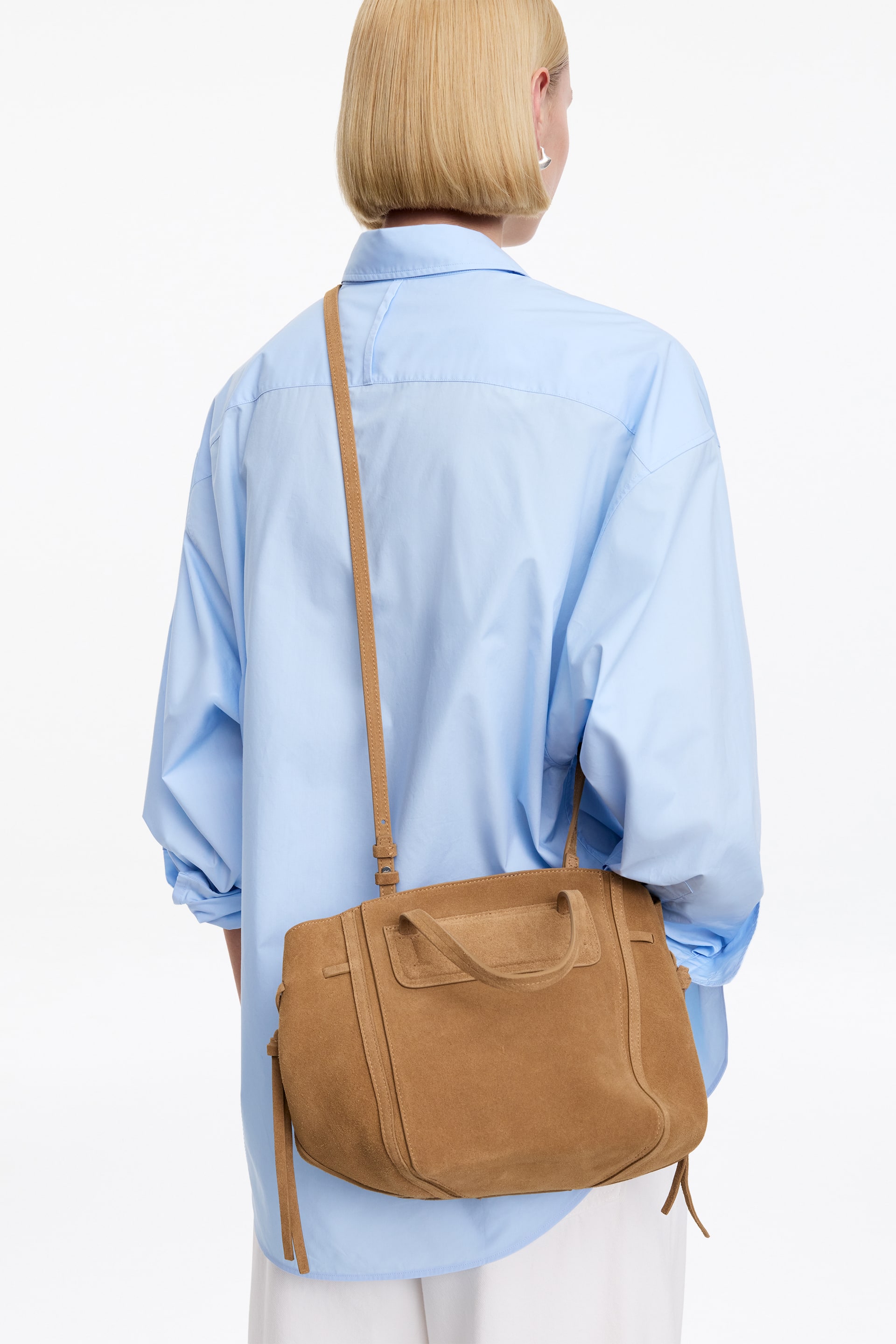 Vis større bilde: En lysebrun skulderveske i semsket skinn henger på en persons høyre side, med fronten vendt mot betrakteren og viser korte topphåndtak, en frontlomme-detalj og sidetrekk med hengende dusker. En justerbar lang stropp strekker seg fra vesken over personens skulder.