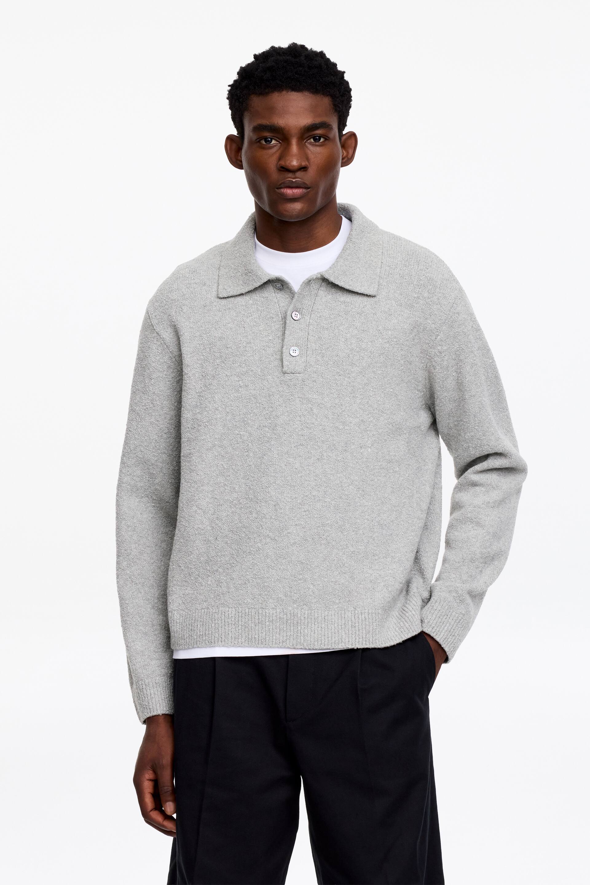 Ver imagem maior: A modelo está a usar uma camisa polo de malha cinza-clara, com gola canelada e meio carcela com três botões de madrepérola. A camisa, em ponto liso com bainha canelada, está sobreposta a uma t-shirt branca e combinada com calças pretas, enquanto a mão da modelo repousa no bolso das calças.