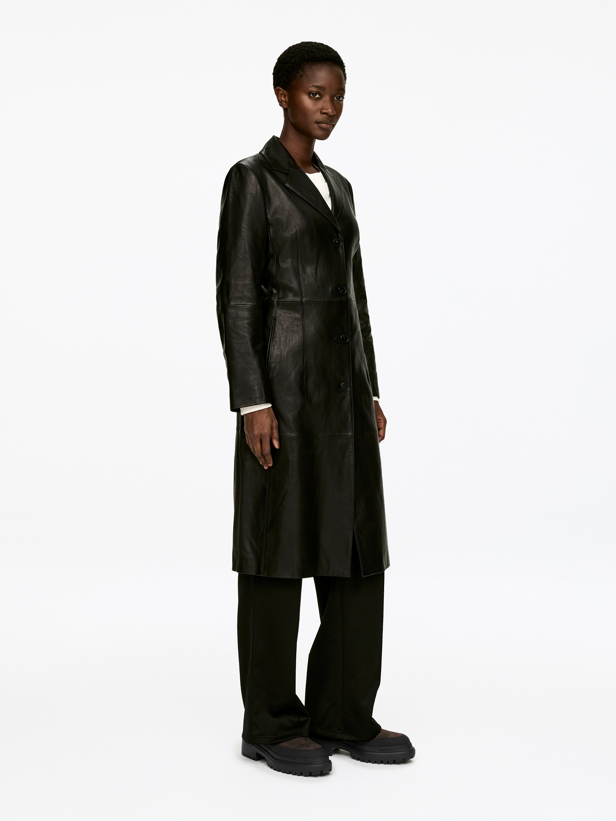 Agrandir l'image: Un manteau en cuir noir est présenté à un léger angle vers la droite. Le manteau de longueur mi-mollet affiche des revers crantés et une fermeture à boutons sur le devant, tandis que sa silhouette droite se superpose à un haut blanc et un pantalon foncé.