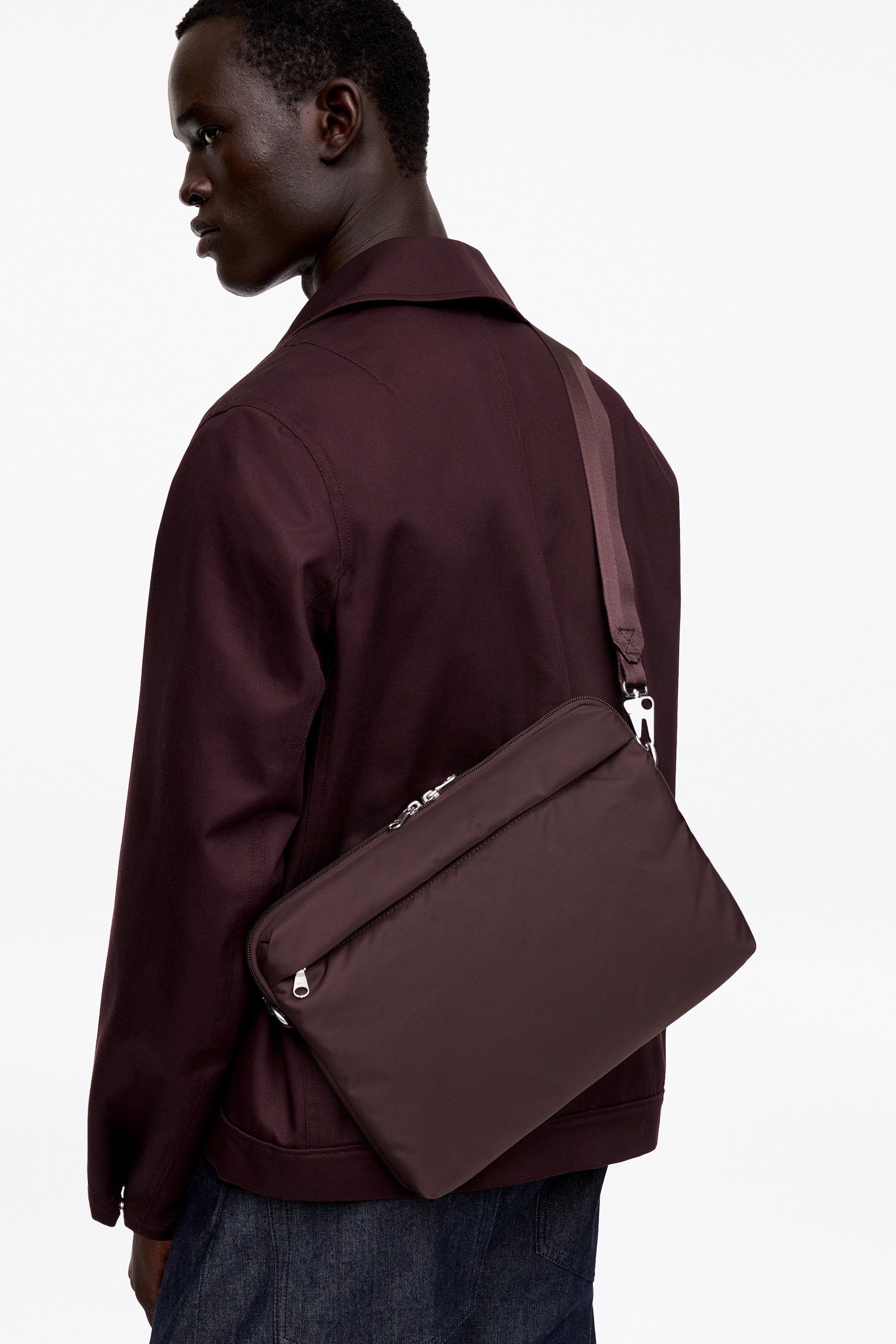 Ver imagen más grande: Un bolso bandolera rectangular de color burdeos oscuro se lleva cruzado sobre el cuerpo, inclinado desde el hombro izquierdo del usuario hasta la cadera derecha. Muestra un cierre principal con cremallera a lo largo de su borde superior, un bolsillo más pequeño con cremallera en la parte delantera y una correa ajustable a juego sujeta con herrajes de tono plateado.