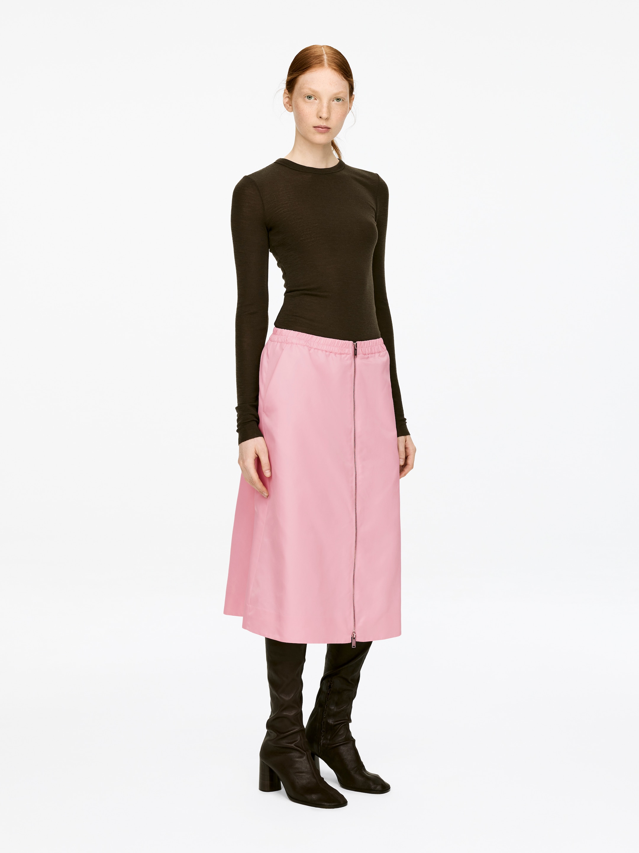 View larger image: A pink midi skirt, featuring an elasticated waist and a two-way zip down the front, is being worn by the model. The skirt is styled with a dark long-sleeved top and dark boots.