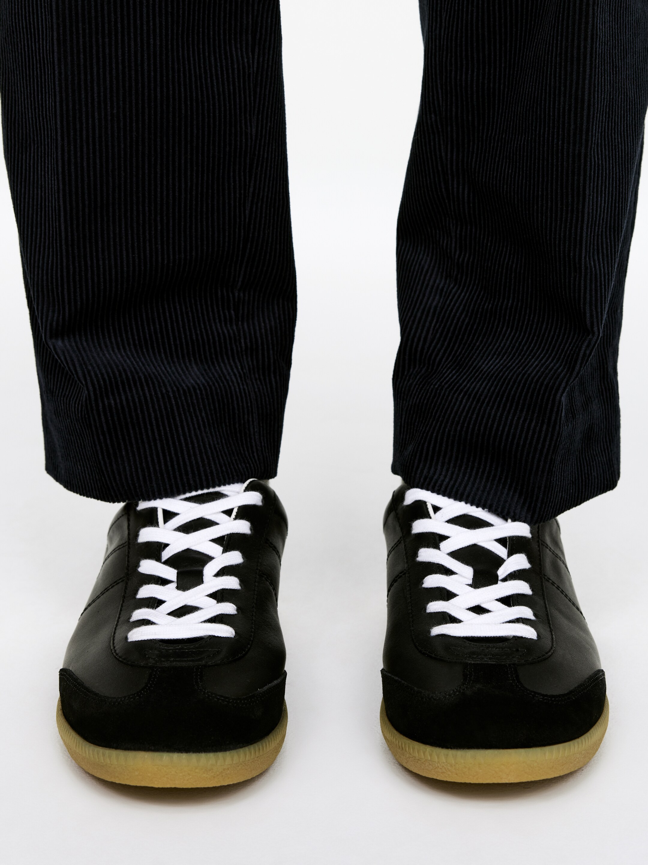 Visualizza immagine più grande: Un paio di sneakers nere basse è indossato, rivolto direttamente in avanti. Le sneakers presentano tomaie in pelle nera con puntali in suede nero, lacci bianchi e suole in gomma marrone chiaro, abbinate a pantaloni in velluto a coste larghe e scure visibili dalla metà del polpaccio in giù.