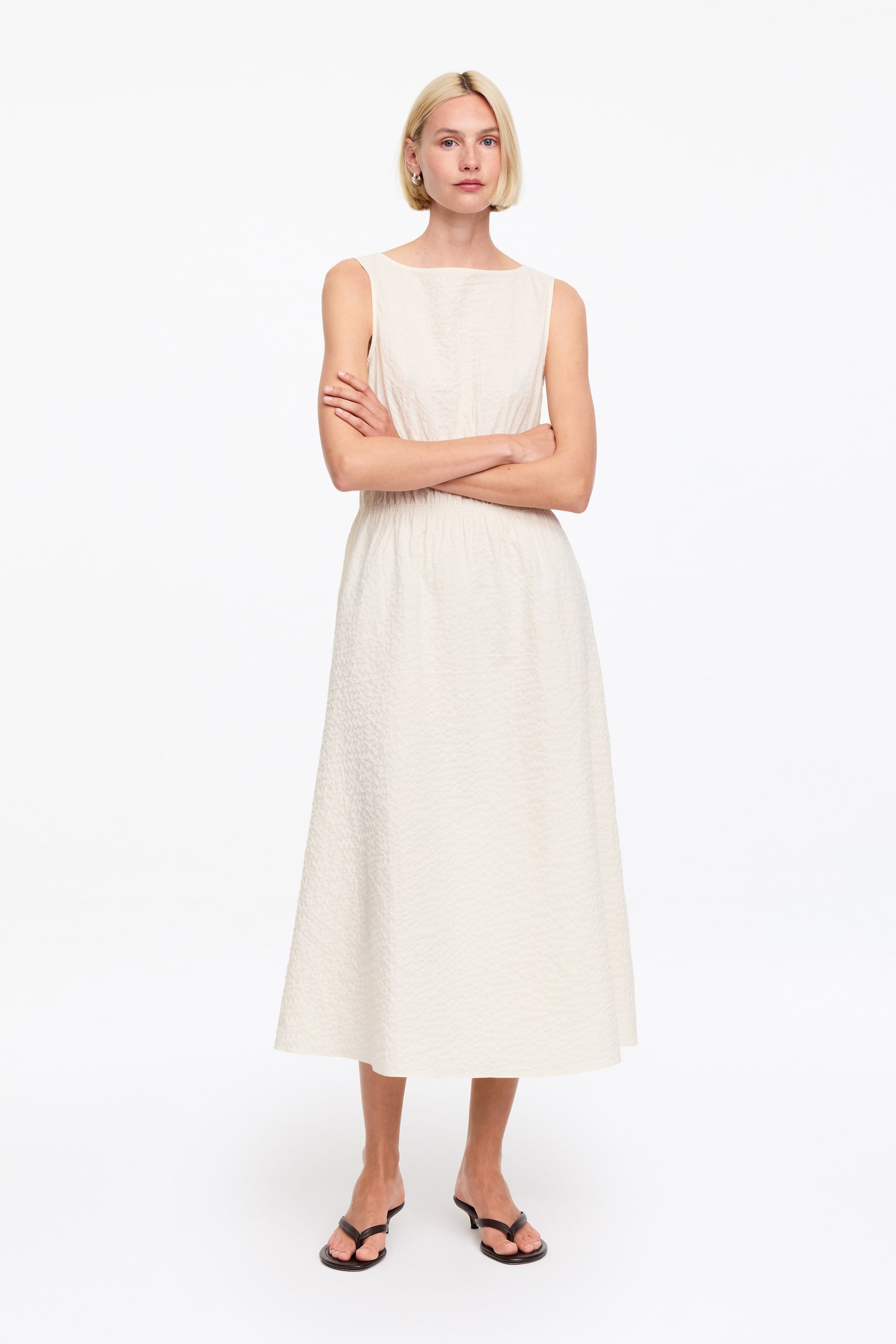 Agrandir l'image: Une robe midi sans manches de couleur crème présente un col bateau, un tissu texturé sur toute la surface, et un corsage ajusté qui se rassemble à la taille avant de s'épanouir en une jupe évasée.