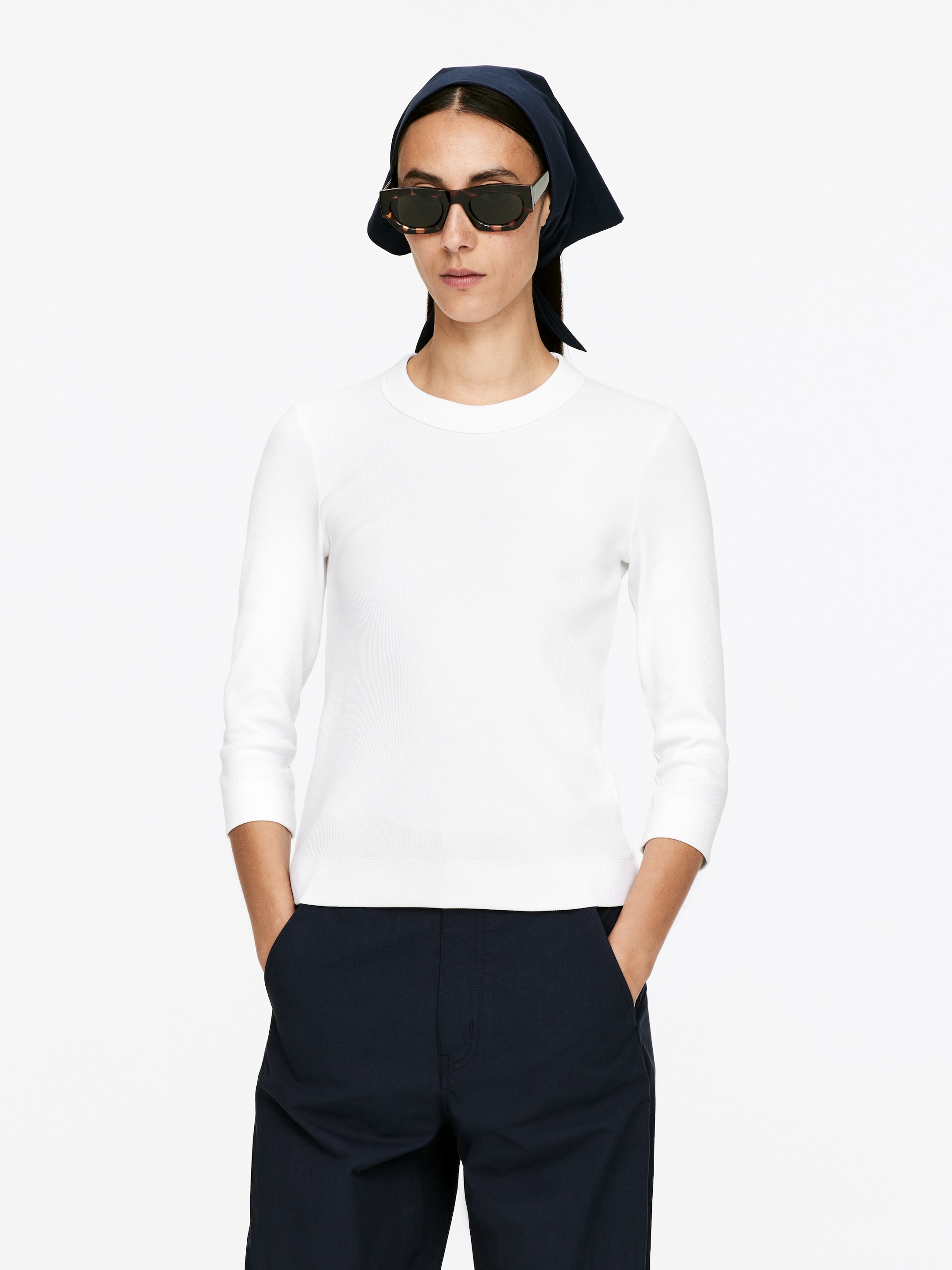 Agrandir l'image: Un haut blanc ajusté à manches trois-quarts est porté, avec un col rond et une texture côtelée subtile. Le haut s'arrête à la taille, où il est associé à un pantalon foncé.