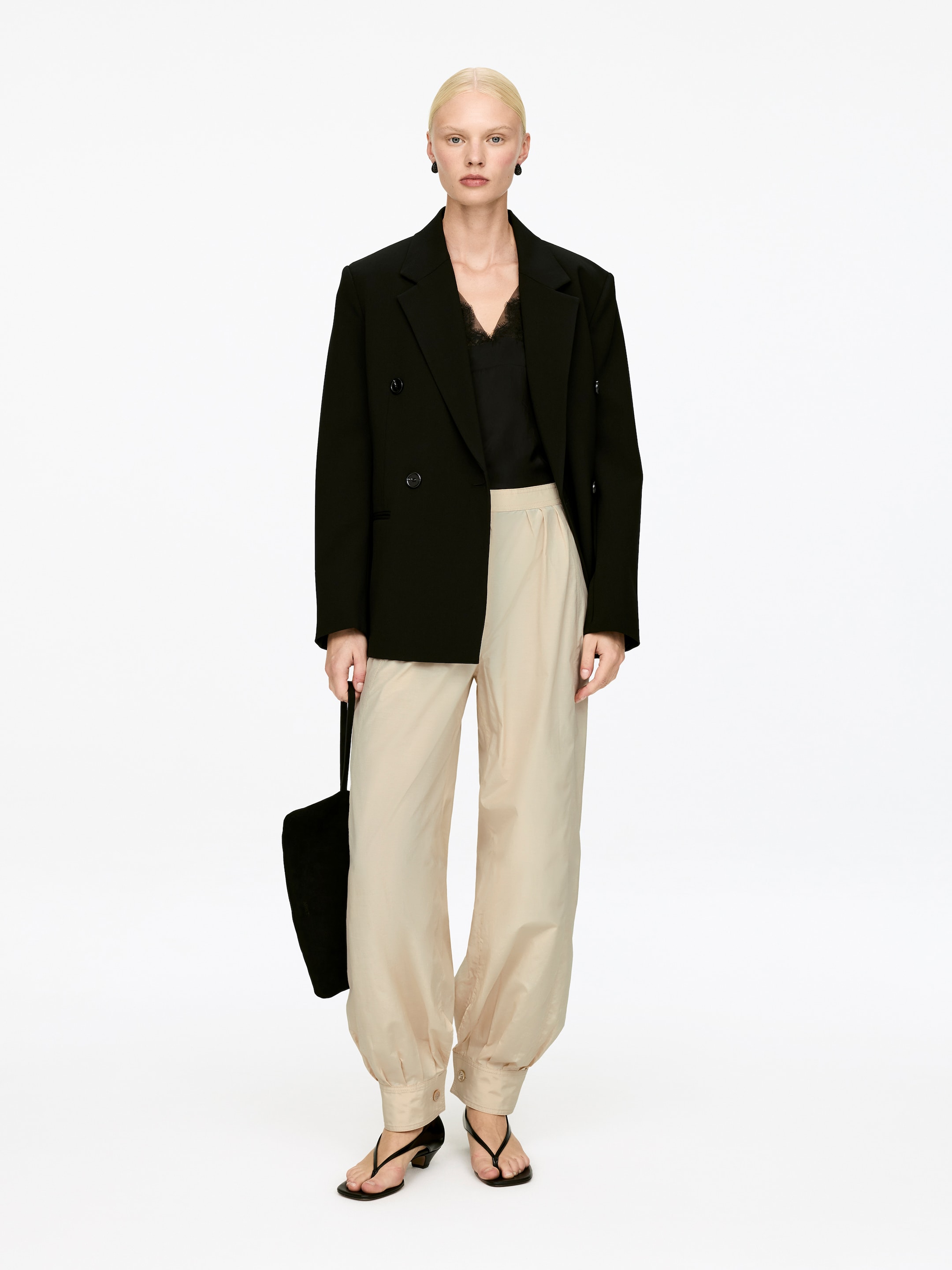 Agrandir l'image: Le mannequin porte un pantalon plissé avec un ourlet bouffant. Le pantalon beige clair est conçu avec des plis visibles à la taille et affiche des boutons sur l'ourlet froncé, qui repose juste au-dessus de sandales noires.