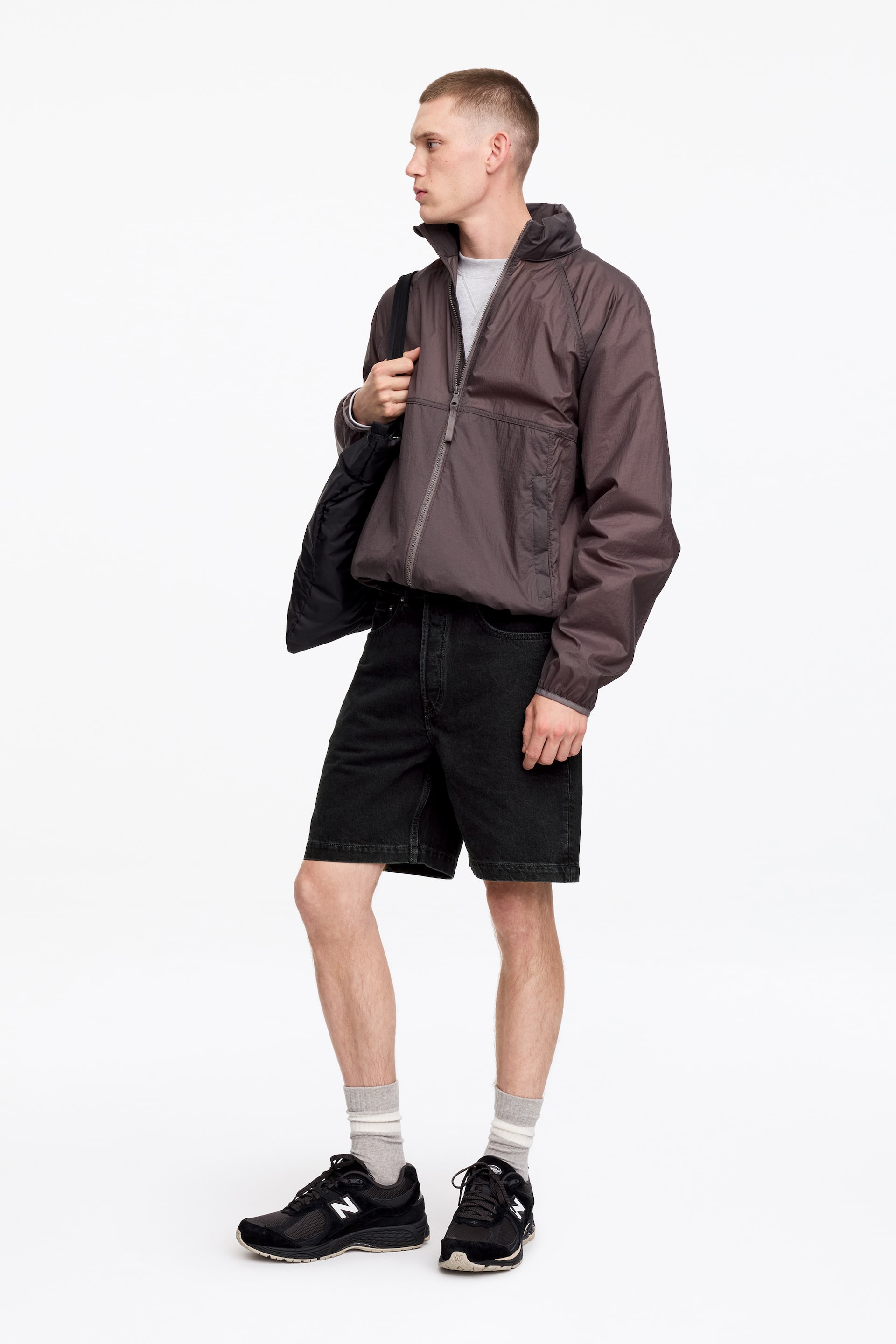 Agrandir l'image: Une veste légère marron foncé est portée vue de côté, affichant une fermeture à demi-zip, un col montant, ainsi que des poignets et un ourlet élastiqués. Elle est stylée par-dessus un t-shirt gris clair, un short en jean noir, des chaussettes rayées grises et des baskets noires.