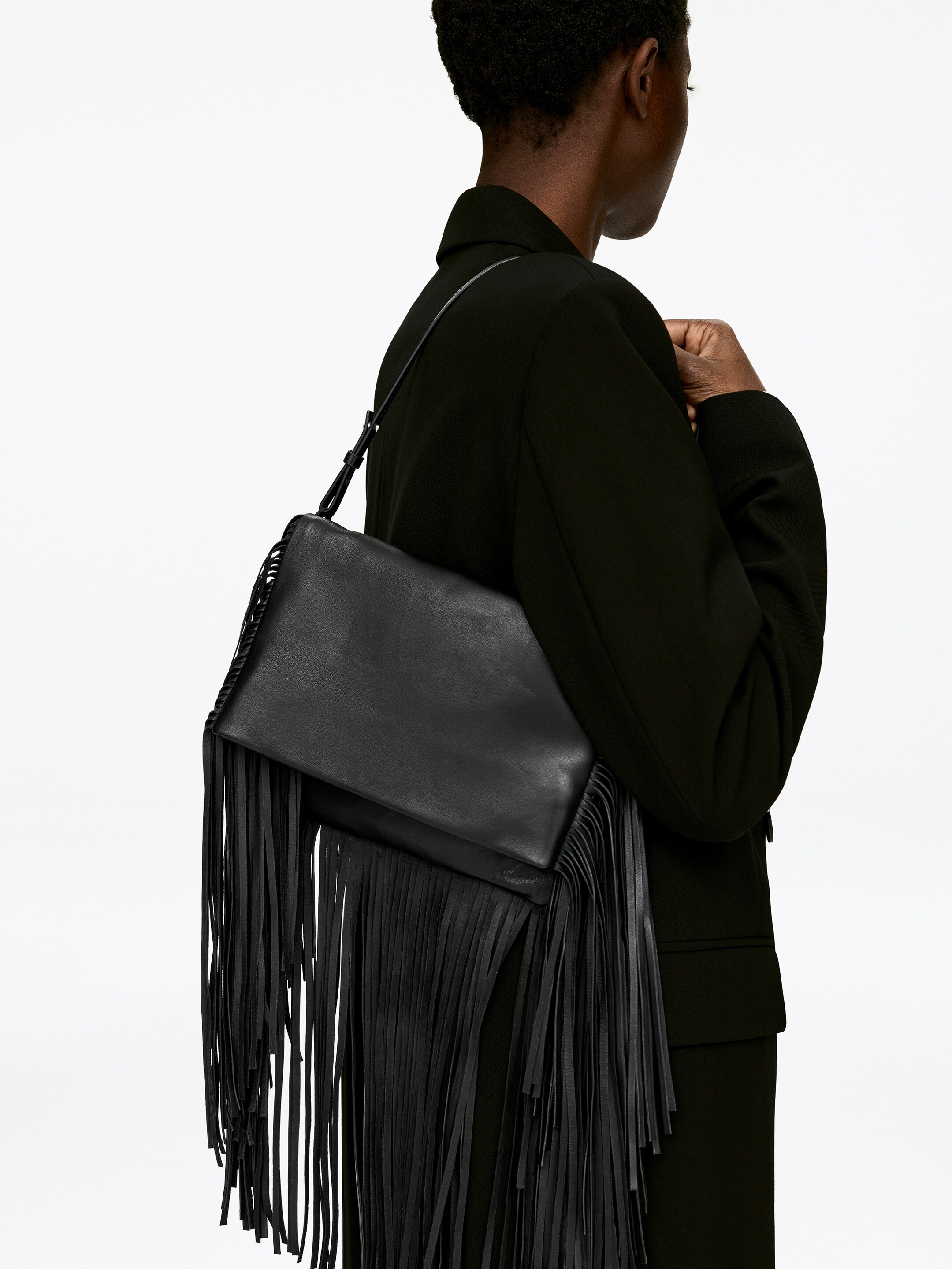 View larger image: A black fringed bag is being presented from a back-right angle, being worn over the shoulder. Its rectangular main section is visible with a smooth flap closure, and long, dense fringe detailing is cascading down its sides and bottom. A thin, adjustable shoulder strap is resting on the shoulder.