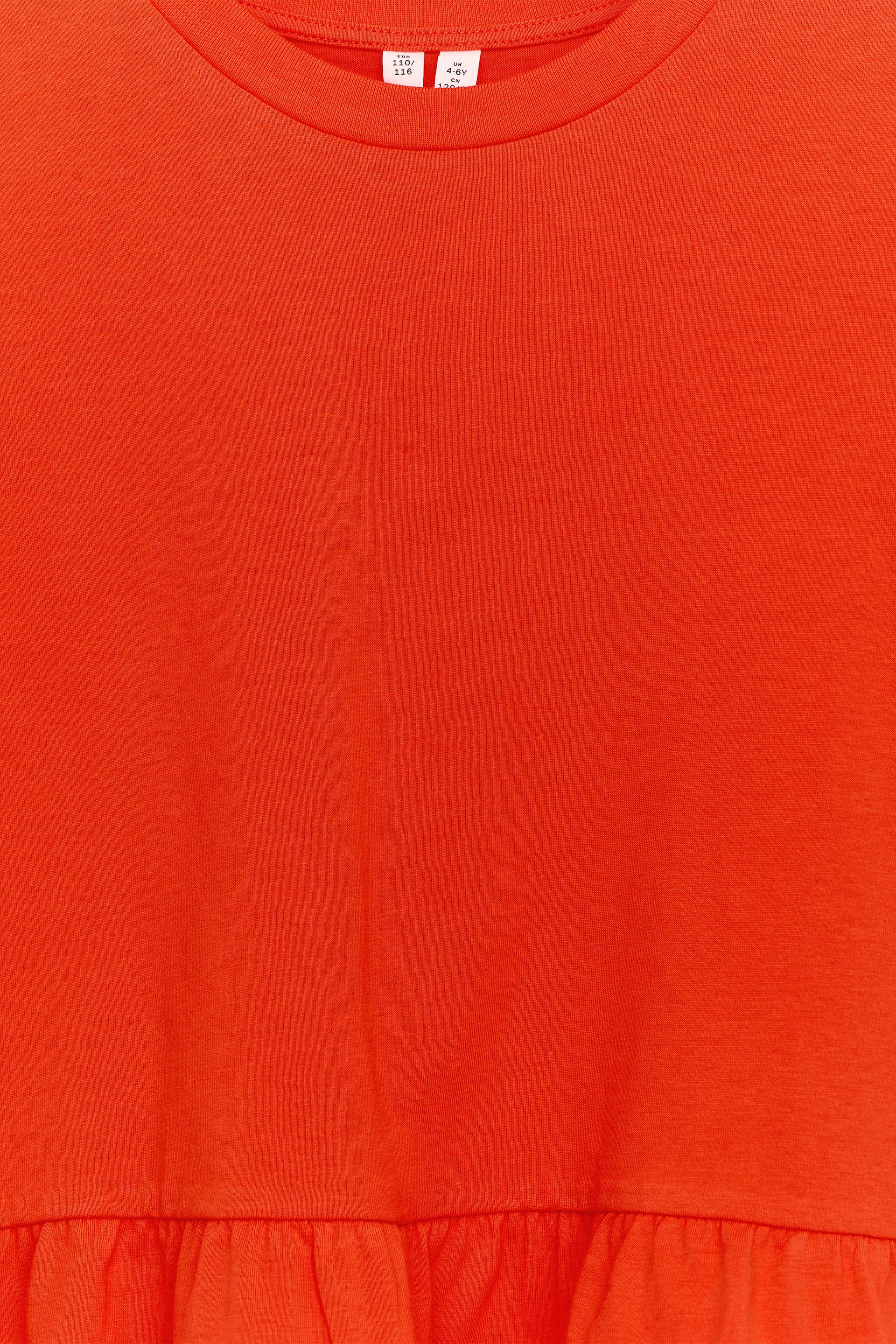 Größeres Bild anzeigen: Ein orange-rotes Kleidungsstück verfügt über einen gerippten Rundhalsausschnitt, mit einem weißen Größenetikett, das oben am Ausschnitt sichtbar ist. Die Vorderseite des Kleidungsstücks zeigt einen glatten, einfarbigen Strickstoff, der in eine geraffte, gerüschte Naht übergeht, die am unteren Ende ein Schößchen-Detail bildet.