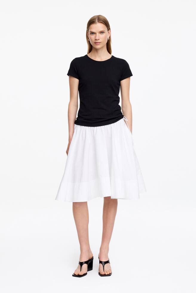 La modelo lleva una falda circular de algodón blanco, que presenta una cintura fruncida con tiras de rouleau atadas al costado y un bolsillo lateral donde descansa la mano de la modelo. La falda cae justo por debajo de la rodilla con una silueta fluida. - 1