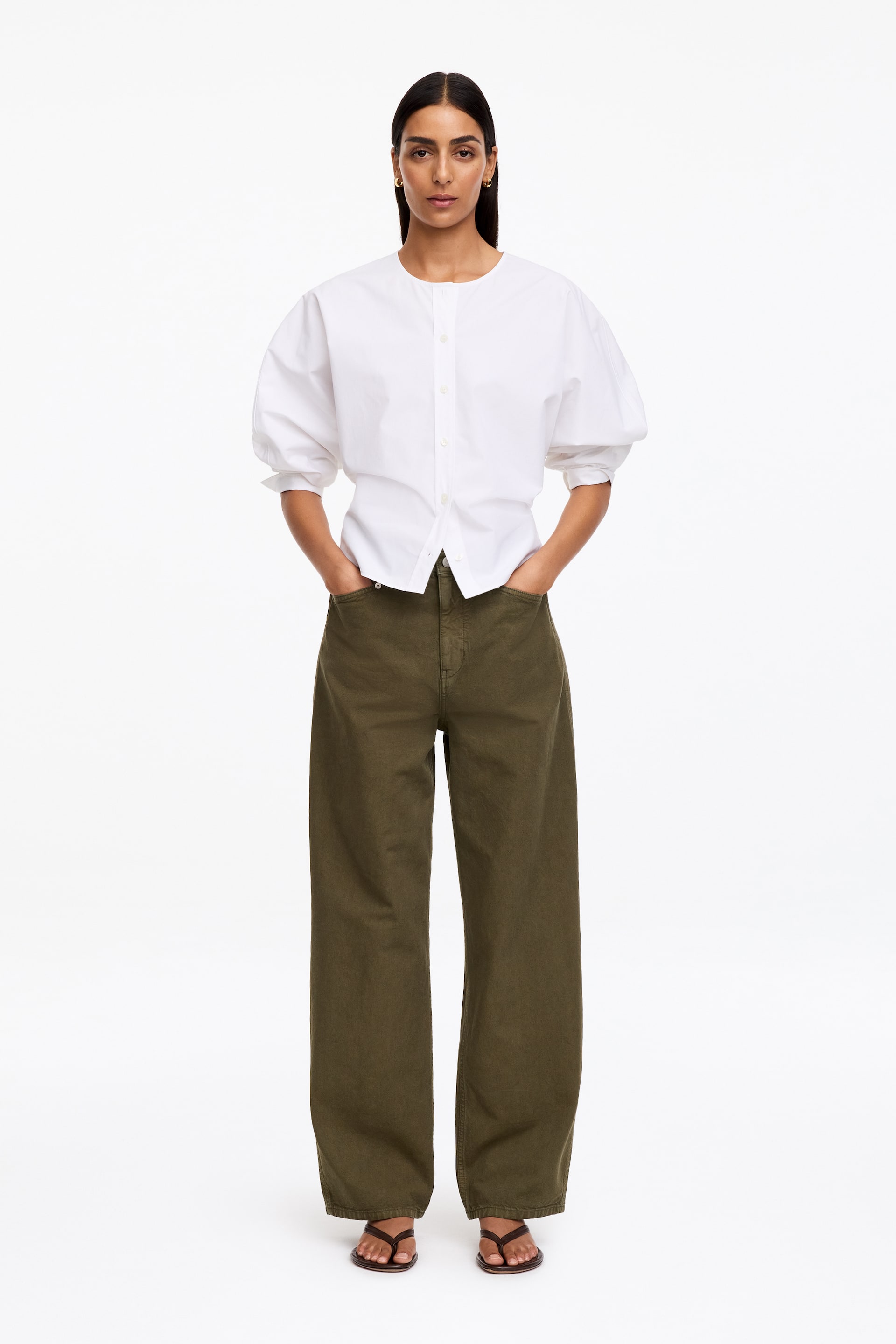 Grotere afbeelding bekijken: Olijfgroene canvas broek, uitgevoerd in een ontspannen, wijde pasvorm met een normale taille, wordt gedragen door het model. De handen van het model rusten in de voorzakken, en een witte blouse en donkere sandalen worden ook bij de broek gedragen.