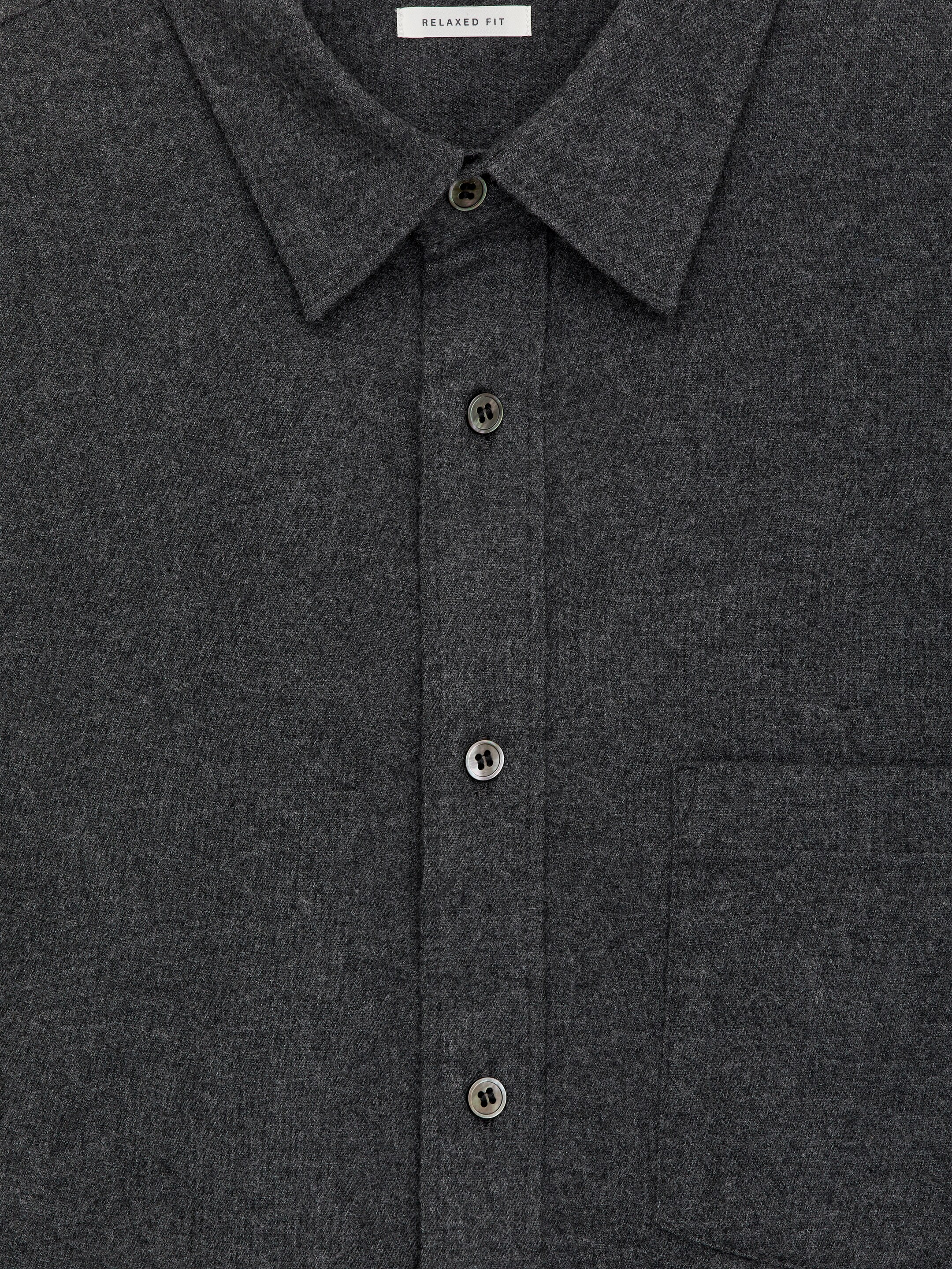 Agrandir l'image: Une chemise en flanelle de coton gris foncé est exposée de face, avec un col standard, une patte de boutonnage à l'avant avec des boutons en nacre, et une poche plaquée sur la poitrine. Une étiquette près du col indique une coupe décontractée.