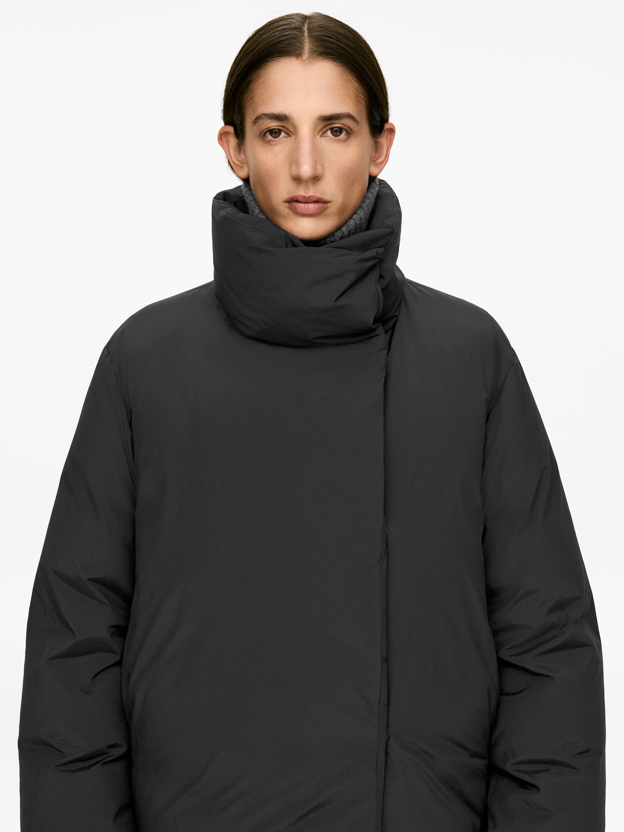 Agrandir l'image: Une veste matelassée gris foncé est portée par le mannequin. La veste se distingue par un col montant structuré et une patte frontale lisse dissimulant des fermetures cachées. Un col roulé gris côtelé est visible dessous le col.
