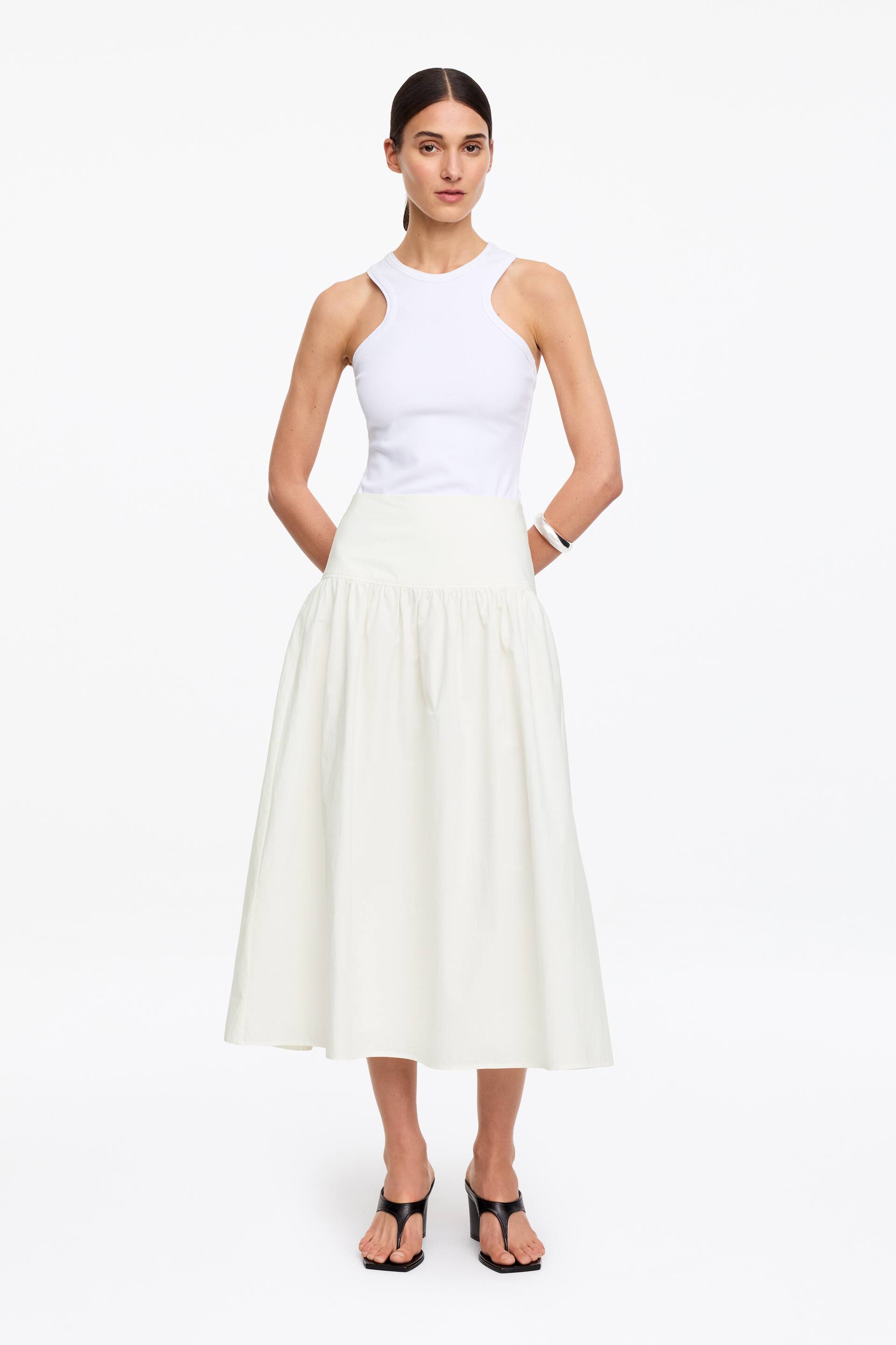 Visualizza immagine più grande: Una gonna lunga color bianco sporco, realizzata in un tessuto seersucker leggermente strutturato, è indossata dalla modella. Questa gonna presenta una fascia in vita ampia e liscia e una silhouette voluminosa e raccolta che cade a metà polpaccio, abbinata a un top bianco a coste infilato dentro.