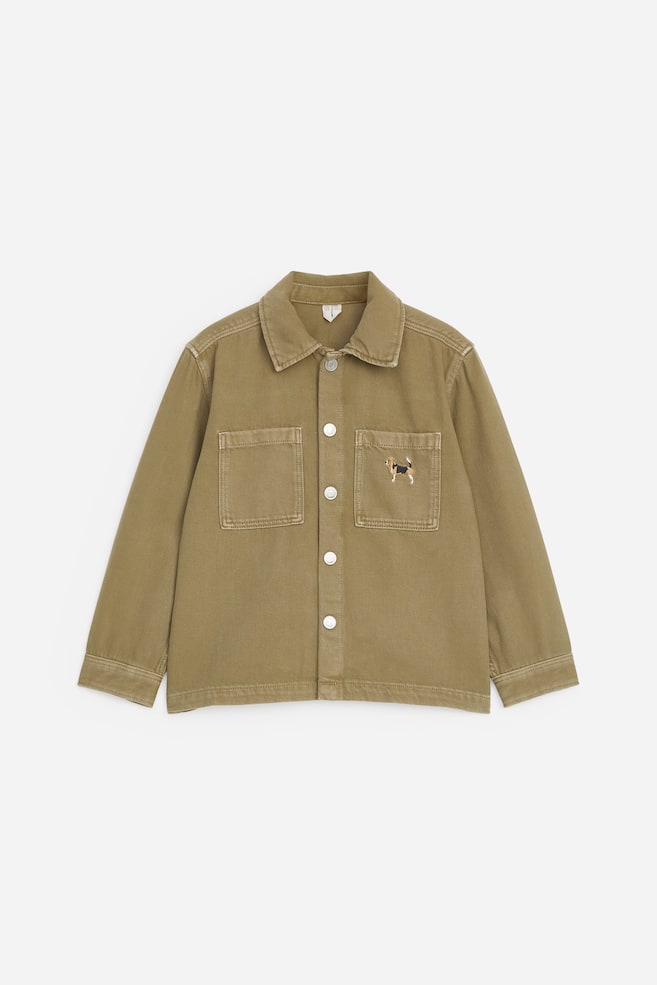 Uma overshirt de ganga verde-khaki é apresentada numa vista frontal plana, com gola clássica, fecho frontal com botões de pressão prateados e mangas compridas com punhos. Dois bolsos de chapa quadrados estão posicionados no peito, sendo que o bolso do lado direito apresenta um pequeno cão bordado. - 1