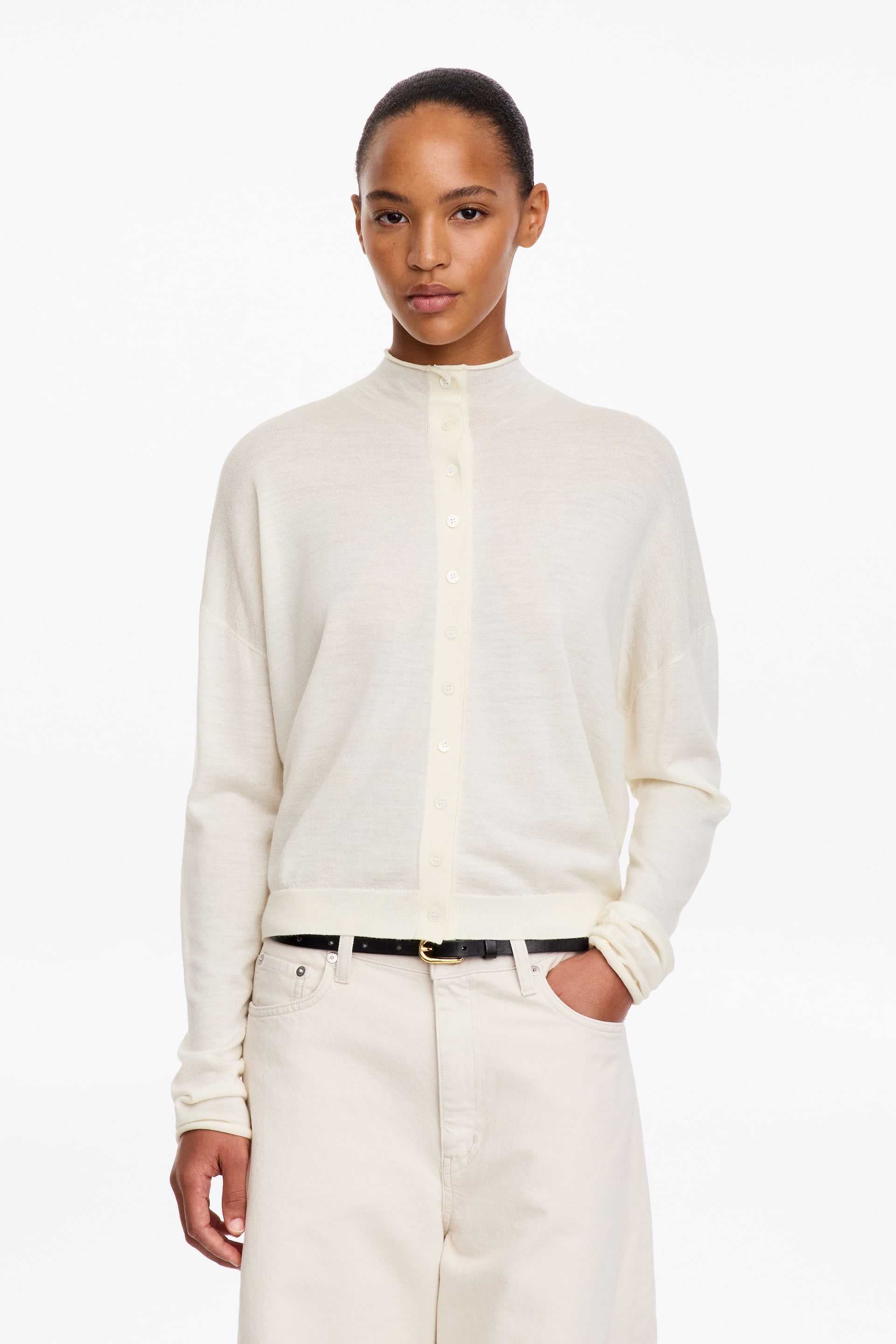 Agrandir l'image: Un cardigan à col montant en mélange de laine, de couleur blanc cassé, est porté par le mannequin. Il présente une fermeture boutonnée sur le devant, des épaules tombantes et des finitions côtelées. Le cardigan est associé à un pantalon crème assorti et à une ceinture noire, le mannequin glissant une main dans une poche.