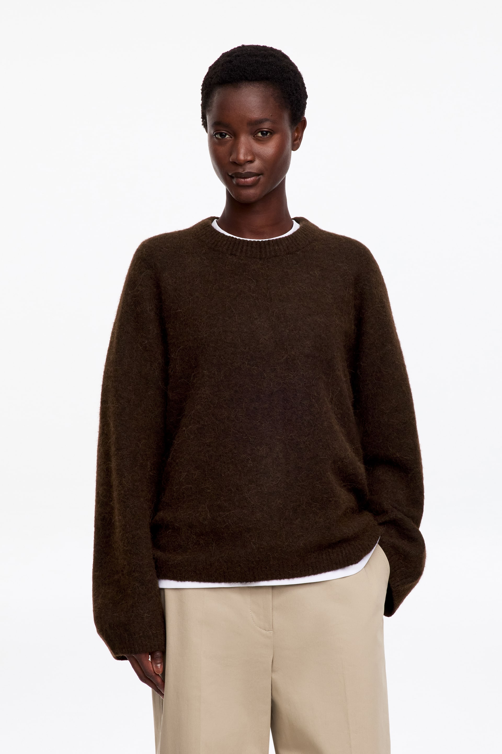 Agrandir l'image: Un pull col rond marron foncé en maille texturée et duveteuse est porté par le mannequin. Le pull est superposé à un t-shirt blanc visible au niveau de l’encolure et de l’ourlet, et il est associé à un pantalon beige.