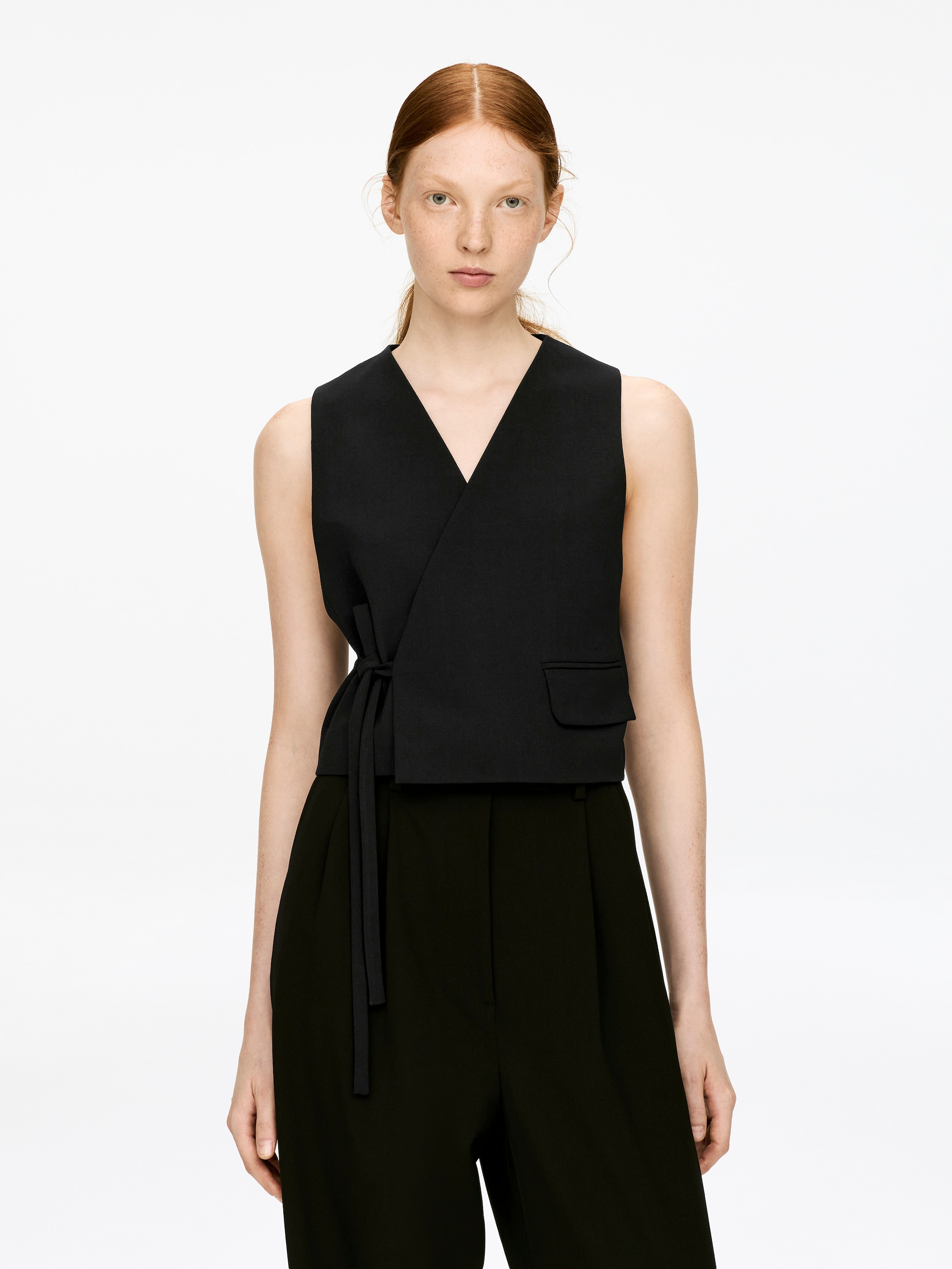 View larger image: A black tailored vest is being worn by the model over dark trousers, featuring an overlapping front secured by a tie strap at the waist, and a decorative pocket flap is visible on the right side.
