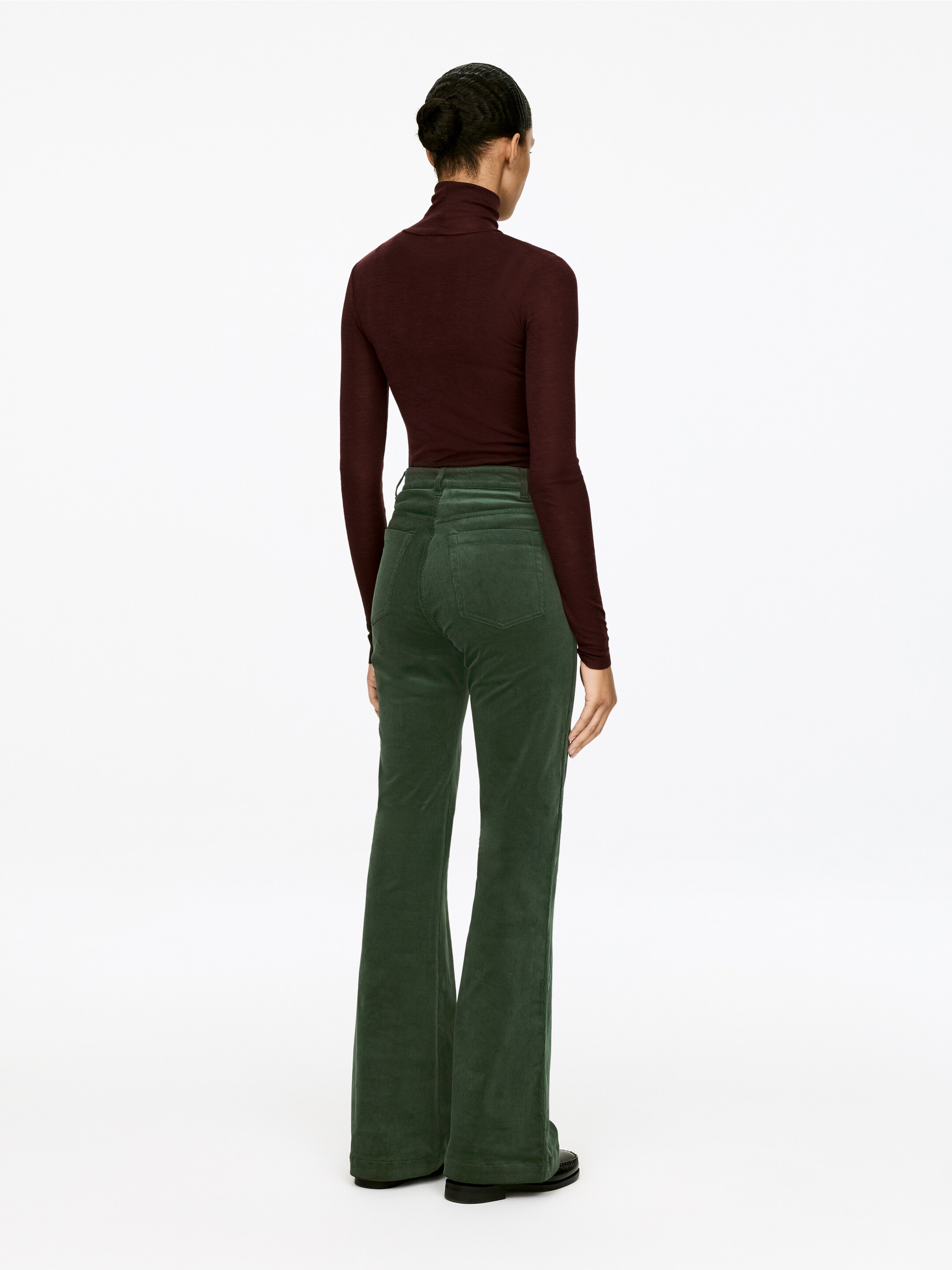 Ver imagen más grande: Pantalones de pana verde oscuro acampanados se presentan desde atrás, con una cintura ajustada y bolsillos traseros. Un top burdeos de cuello alto de manga larga se lleva metido dentro de los pantalones.