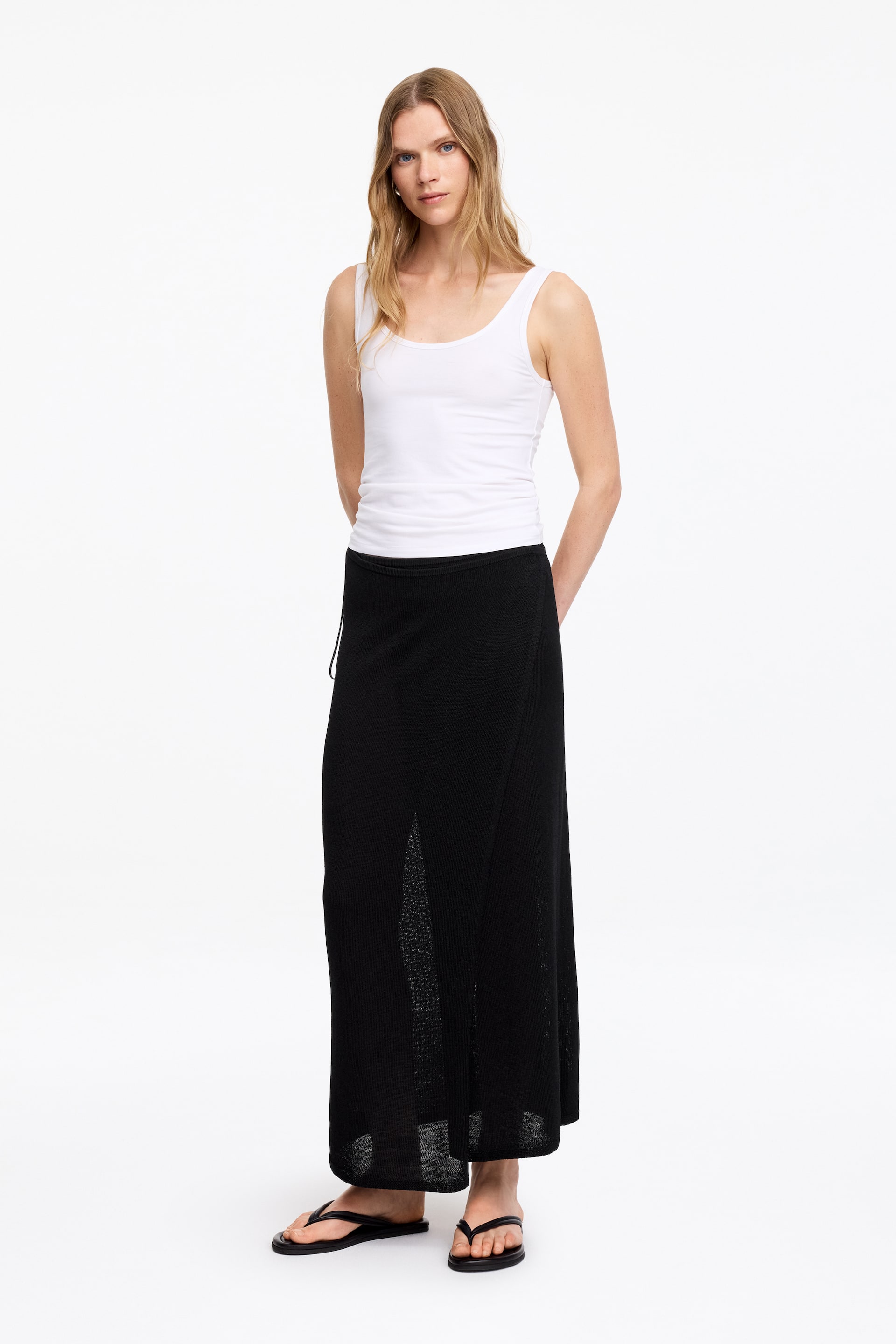 Ver imagen más grande: Una falda maxi de punto negro exhibe un diseño estilo envolvente con una abertura frontal. Presenta una cintura con cordón ajustable y un tejido ligeramente translúcido, visible especialmente en la parte inferior y la abertura.
