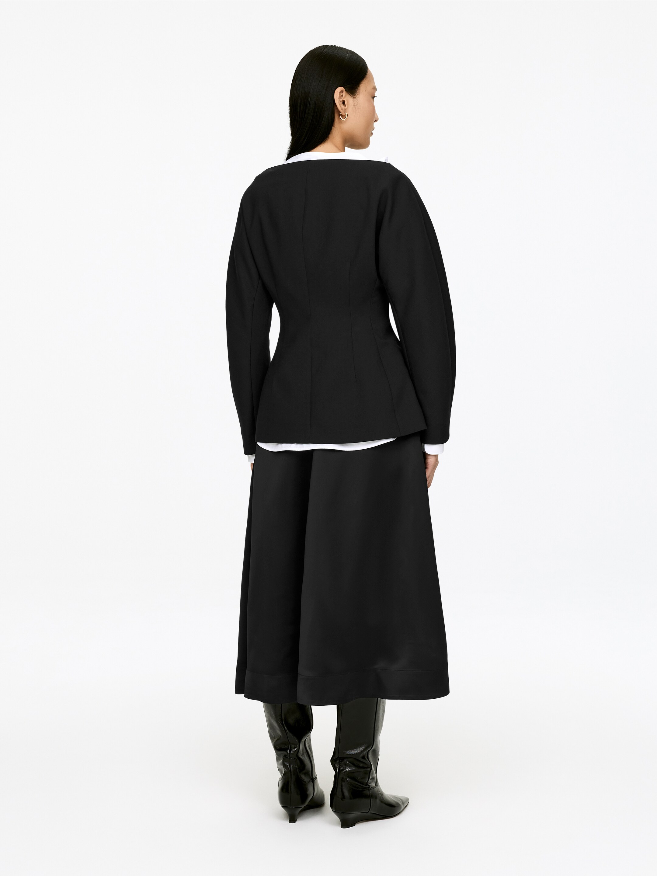 Grösseres Bild ansehen: Eine schwarze, strukturierte Jacke und ein schwarzer, mittellanger ausgestellter Rock werden von hinten präsentiert. Die Jacke hat eine taillierte Taille und Ärmel mit subtilem Volumen und wird über einem weißen Hemd getragen, während der Rock knapp über schwarzen Stiefeln mit spitzer Zehe endet.