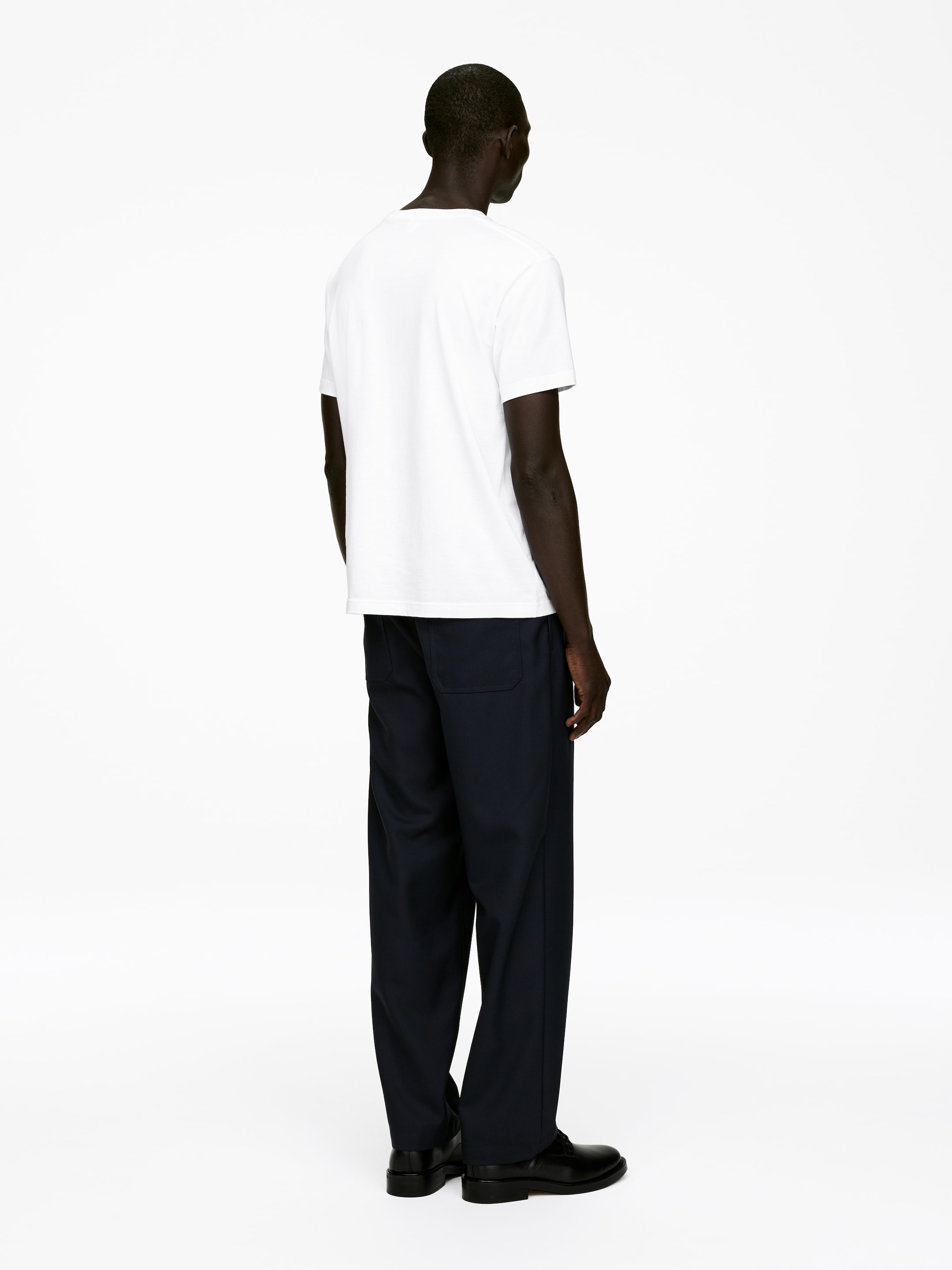 Visa större bild: En vit t-shirt, marinblå byxor och svarta skor visas från en bakre vy. Den vita t-shirten har en avslappnad passform med korta ärmar och en enkel rund halsringning. De marinblå byxorna visar en bredbent silhuett, en elastisk midjeband och synliga bakfickor, med svarta läderskor som också bärs.