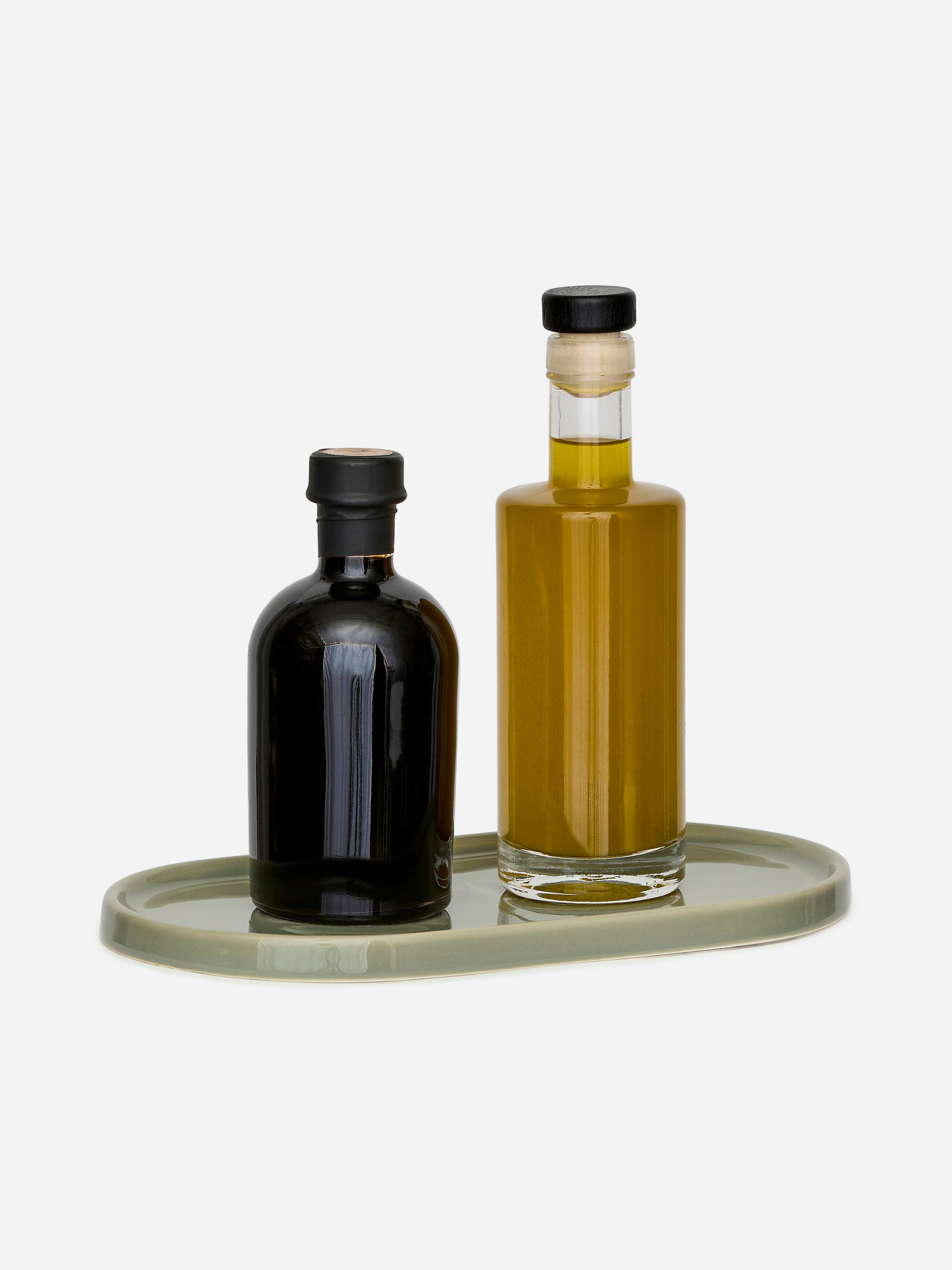 Visualizza immagine più grande: Un piatto in terracotta smaltata ha una forma ovale, presenta un bordo rialzato poco profondo ed è di un verde salvia chiaro. Attualmente contiene due bottiglie, una scura e una riempita con olio d'oliva, dimostrando la sua funzione come vassoio.