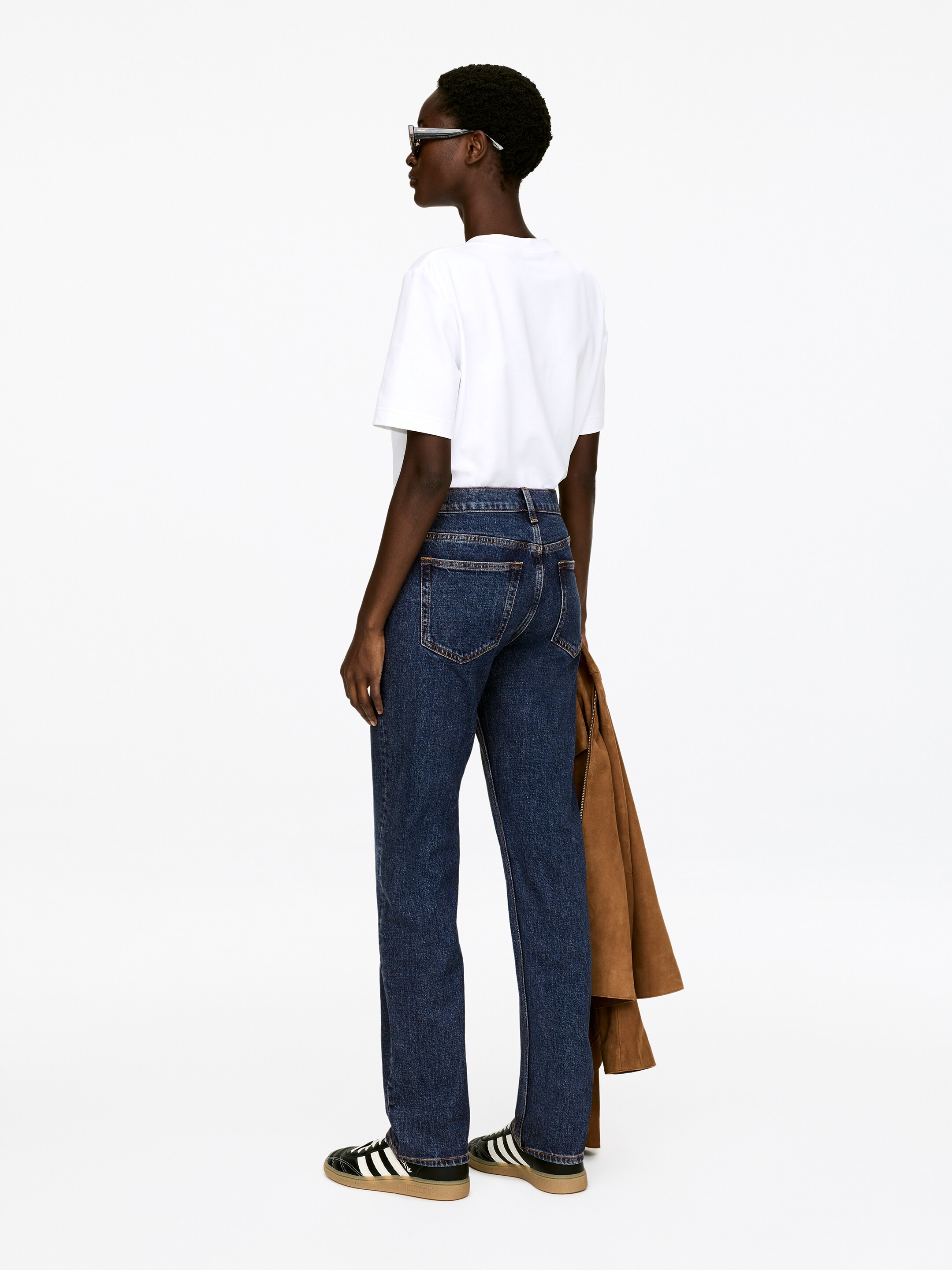 Visualizza immagine più grande: Jeans a gamba dritta con lavaggio scuro sono presentati da una vista posteriore, mostrando tasche posteriori e passanti per la cintura visibili. Una maglietta bianca con scollo a girocollo è indossata e una giacca marrone è portata sopra il braccio.