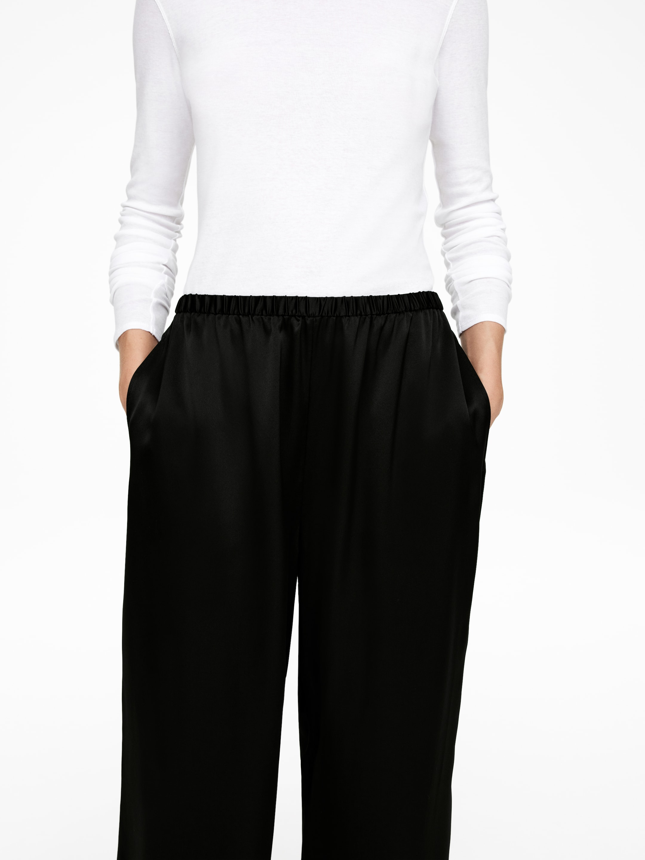 View larger image: Black satin-finish pants are featuring an elasticated, gathered waistband and side pockets. Hands are visibly resting inside the pockets, showcasing the pants' relaxed, wide-leg silhouette.