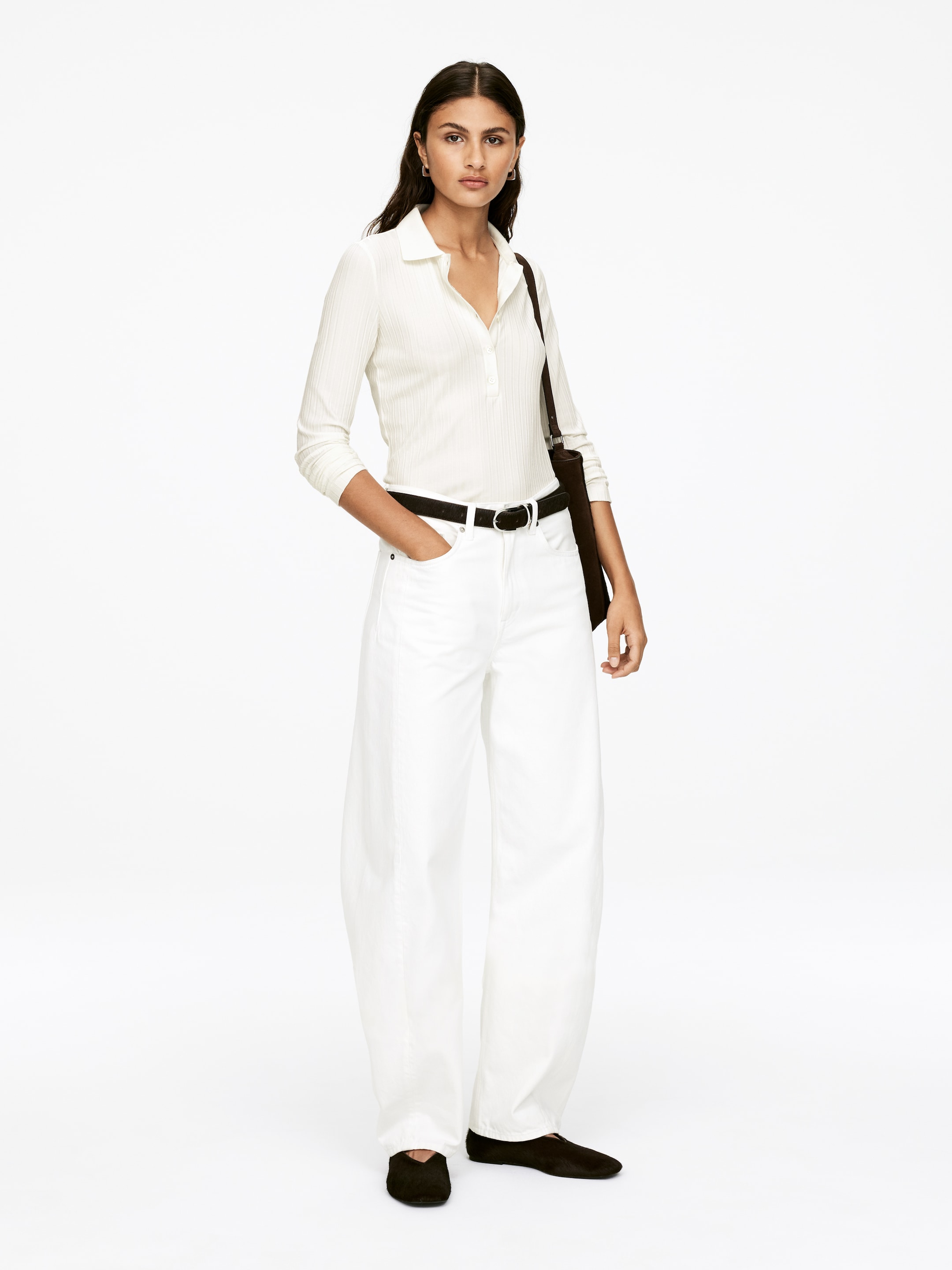 Agrandir l'image: Un polo blanc ajusté en maille côtelée avec une patte de boutonnage et des manches longues est présenté, rentré dans un pantalon blanc à taille haute et à jambes larges. Le pantalon est serré avec une ceinture noire et tombe légèrement sur les chevilles.
