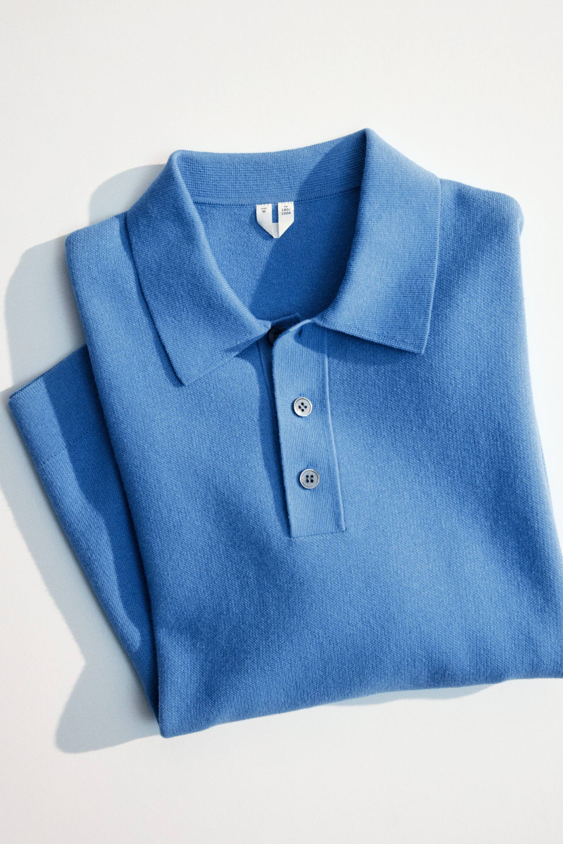 Visualizza l'immagine ingrandita: Una maglia polo blu in maglia è stesa piatta e piegata, con un colletto classico e una chiusura a due bottoni con bottoni grigio chiaro.