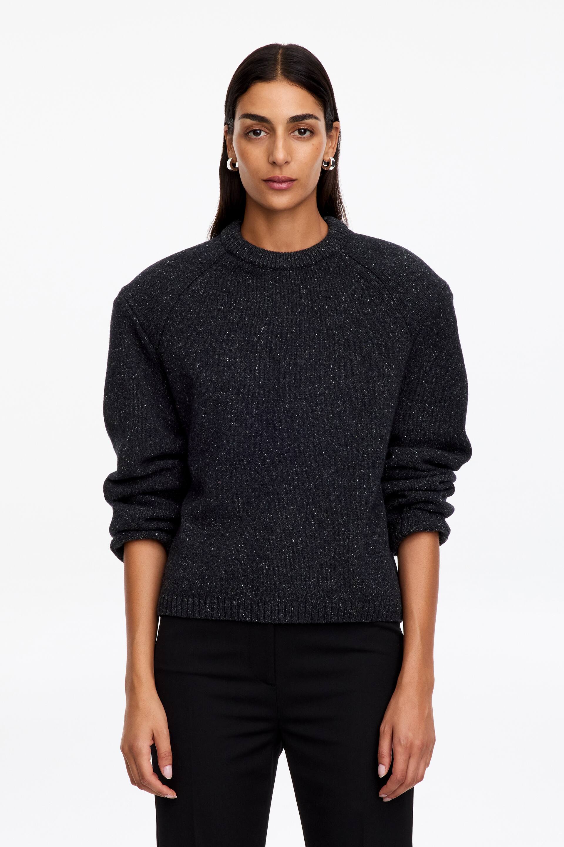 Agrandir l'image: Un pull en maille chinée gris foncé est porté par le modèle, avec un col rond côtelé, des épaules structurées et des manches raglan. Le pull est associé à un pantalon noir, ses manches longues remontées jusqu’au milieu des avant-bras.