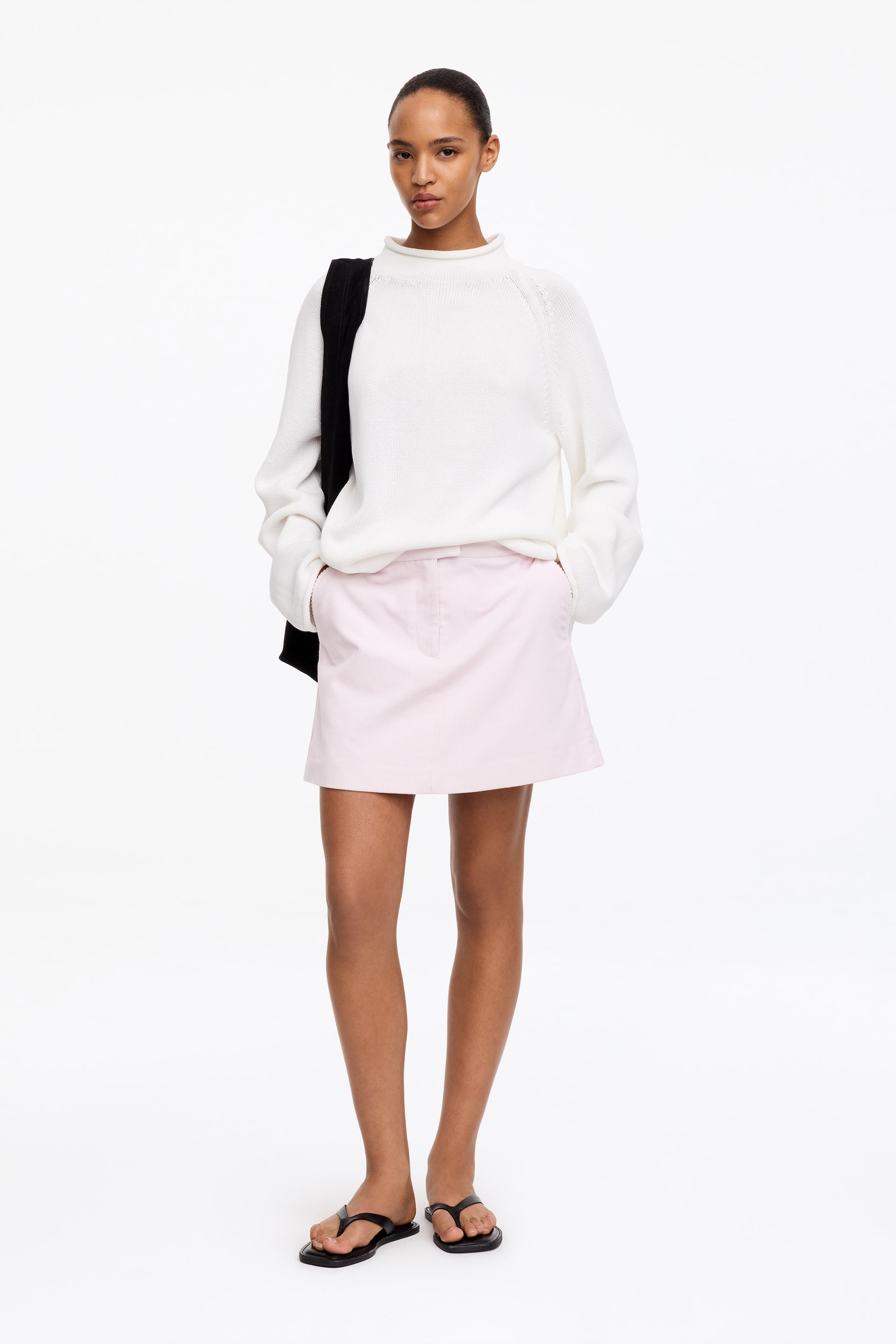 Agrandir l'image: Mini-jupe en velours côtelé. La mini-jupe en velours côtelé à fines côtes, de couleur rose clair, est portée par le mannequin, qui se tient debout les mains dans les poches avant. Elle est associée à un pull en maille blanc.
