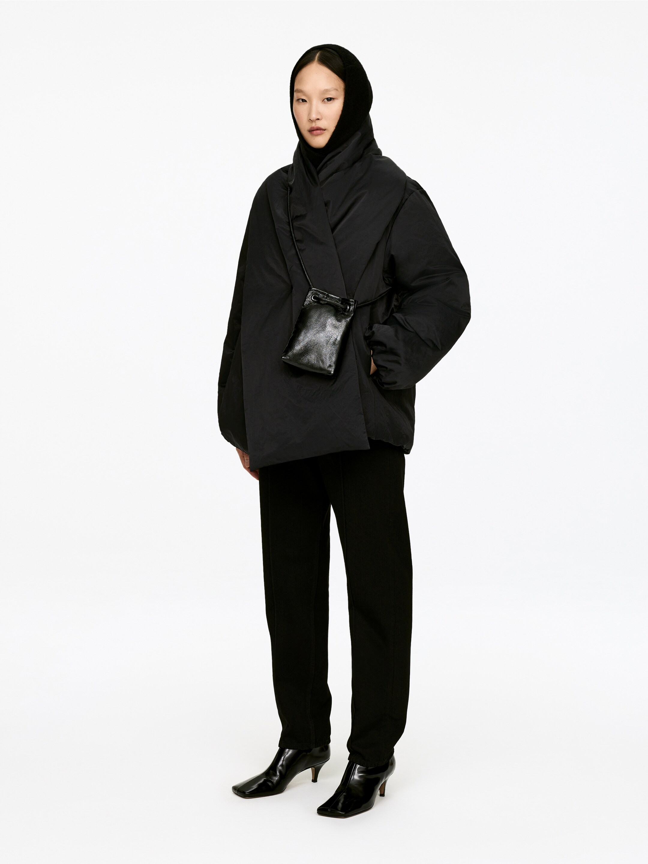 Vis større bilde: En svart vattert wrap-front kåpe vender fremover, med et voluminøst, oversized design med en bred, brettet krage og lange, vide ermer. En liten svart lærveske bæres over kåpen.