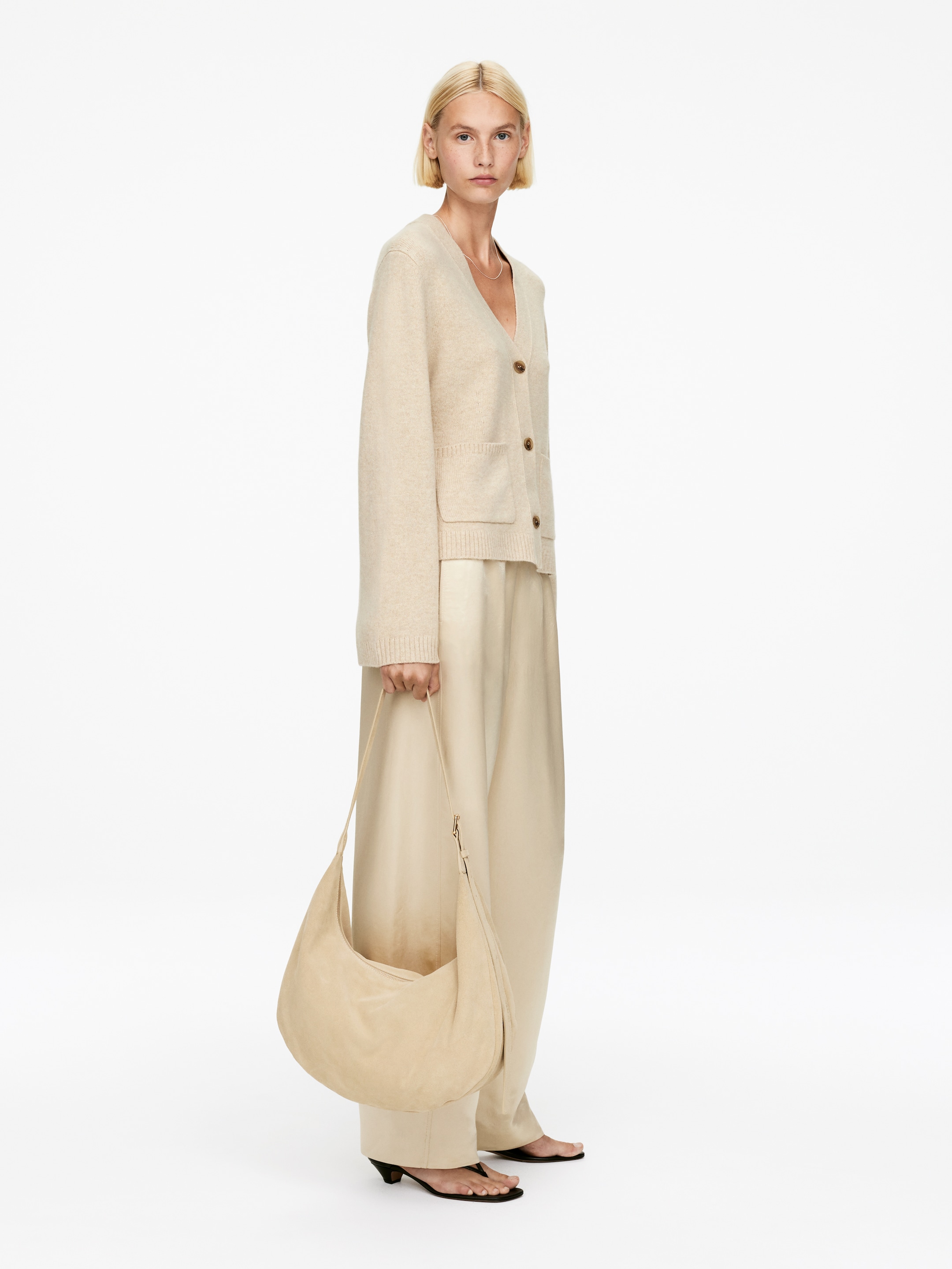 Agrandir l'image: Un cardigan en tricot beige est présenté sous un angle légèrement avant-droit, mettant en valeur sa coupe décontractée. Il présente un décolleté en V, trois boutons marron et deux poches plaquées, avec des poignets et un ourlet côtelés.