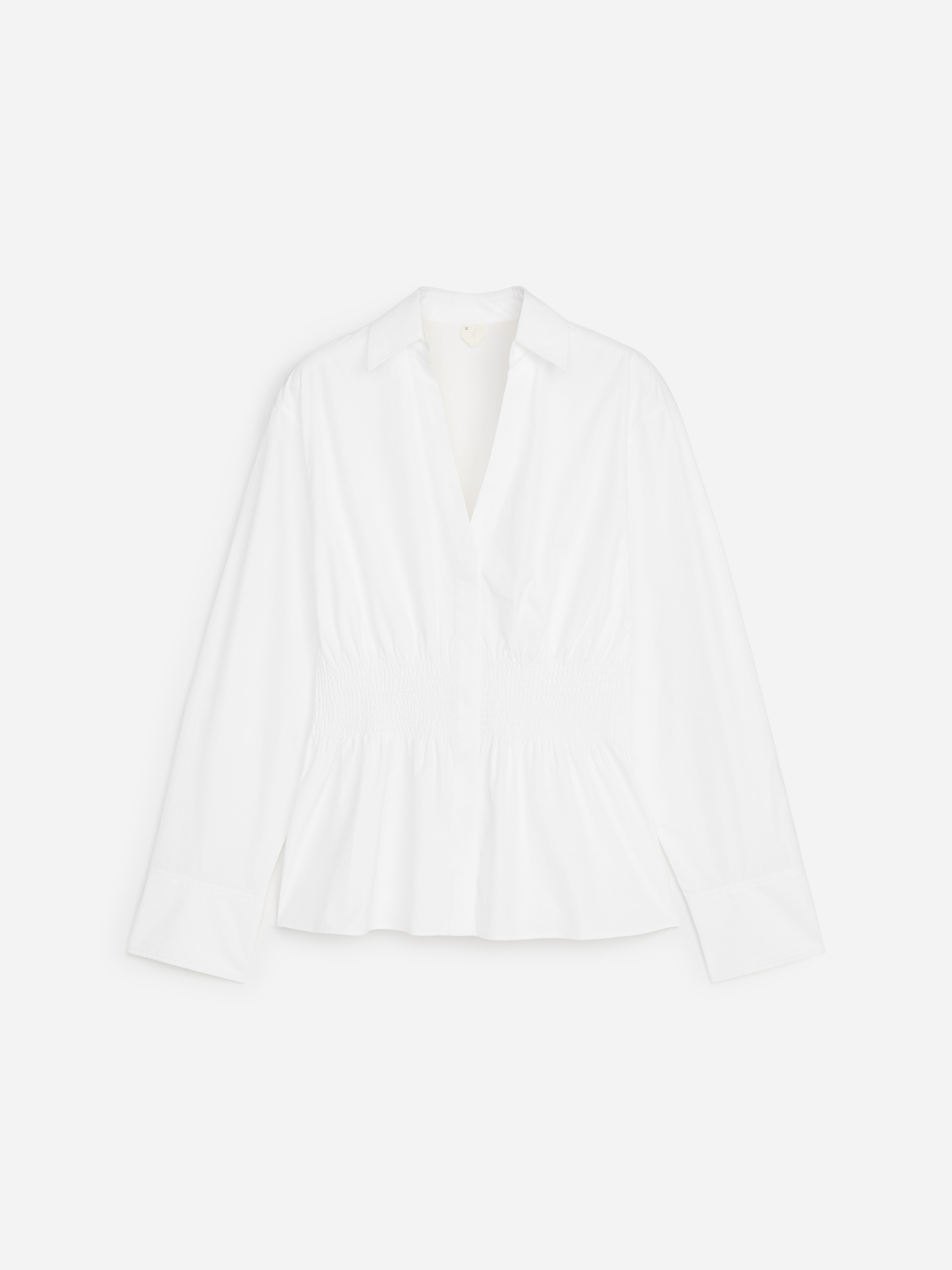 Le mannequin porte une chemise blanche Ã  manches longues, dotÃ©e dâun col en V, de larges poignets et dâune taille cintrÃ©e smockÃ©e. La chemise sâÃ©vase en ourlet style peplum et est associÃ©e Ã  un jean bleu foncÃ©.