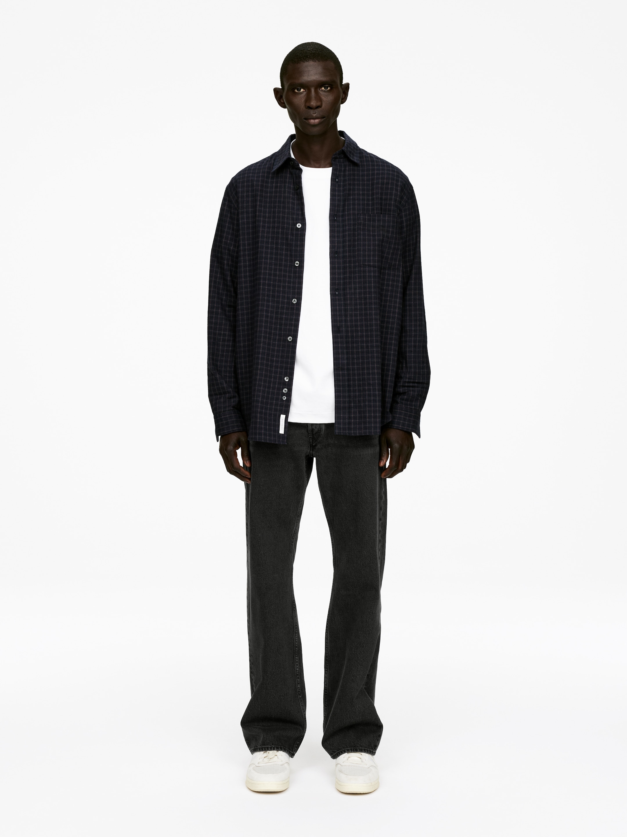 Ingrandisci l'immagine: Una camicia a maniche lunghe blu scuro a quadri viene indossata sbottonata sopra una t-shirt bianca a girocollo, insieme a jeans grigio scuro a vestibilità ampia e sneakers bianche con lacci, il tutto presentato da una vista frontale completa.