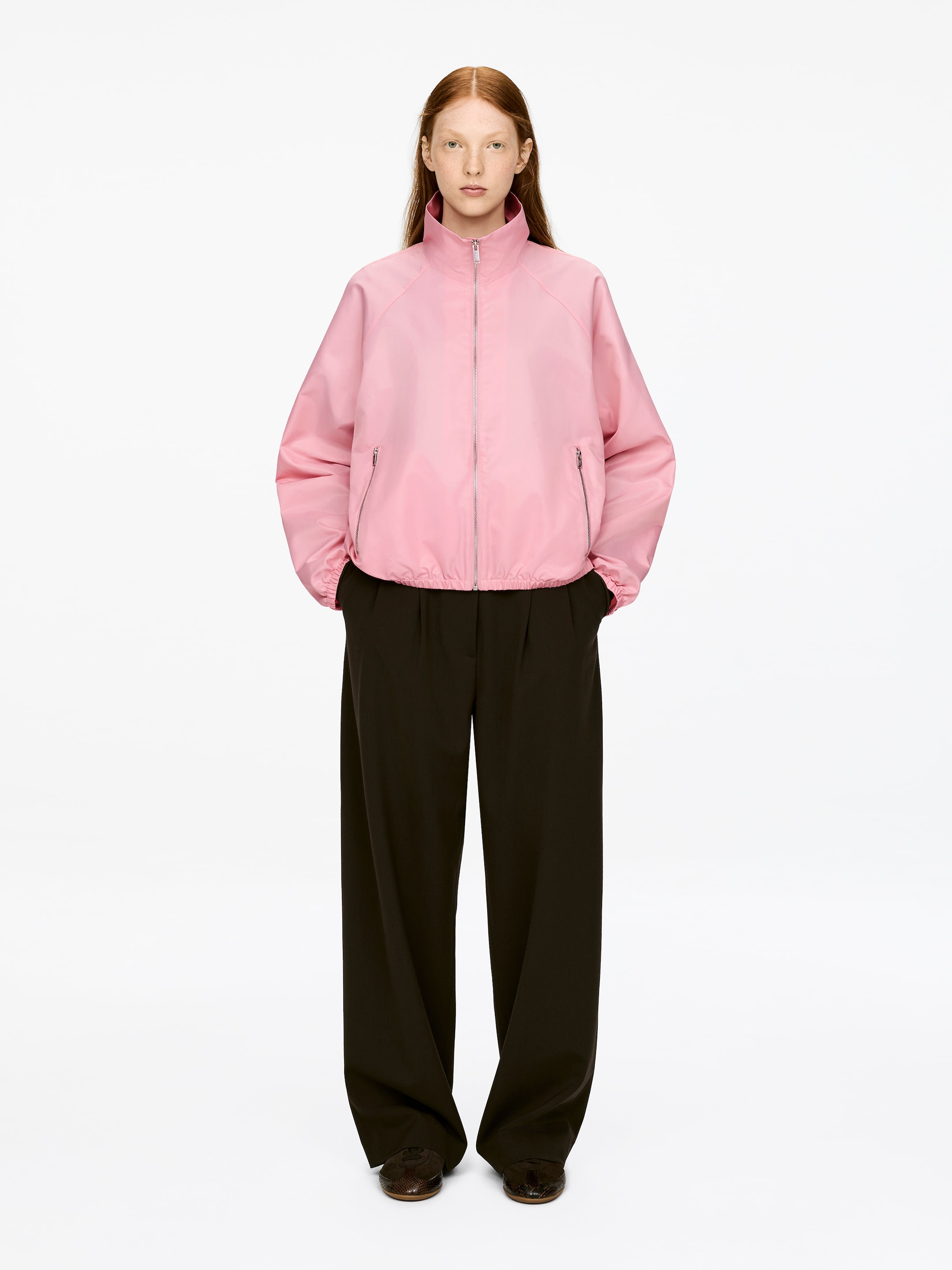 Vis større bilde: En rosa sporty nylonjakke, med en metallglidelås foran, to metallglidelåslommer og elastiske mansjetter og nedre kant, bæres av modellen. Modellen står med hendene i jakkens lommer.