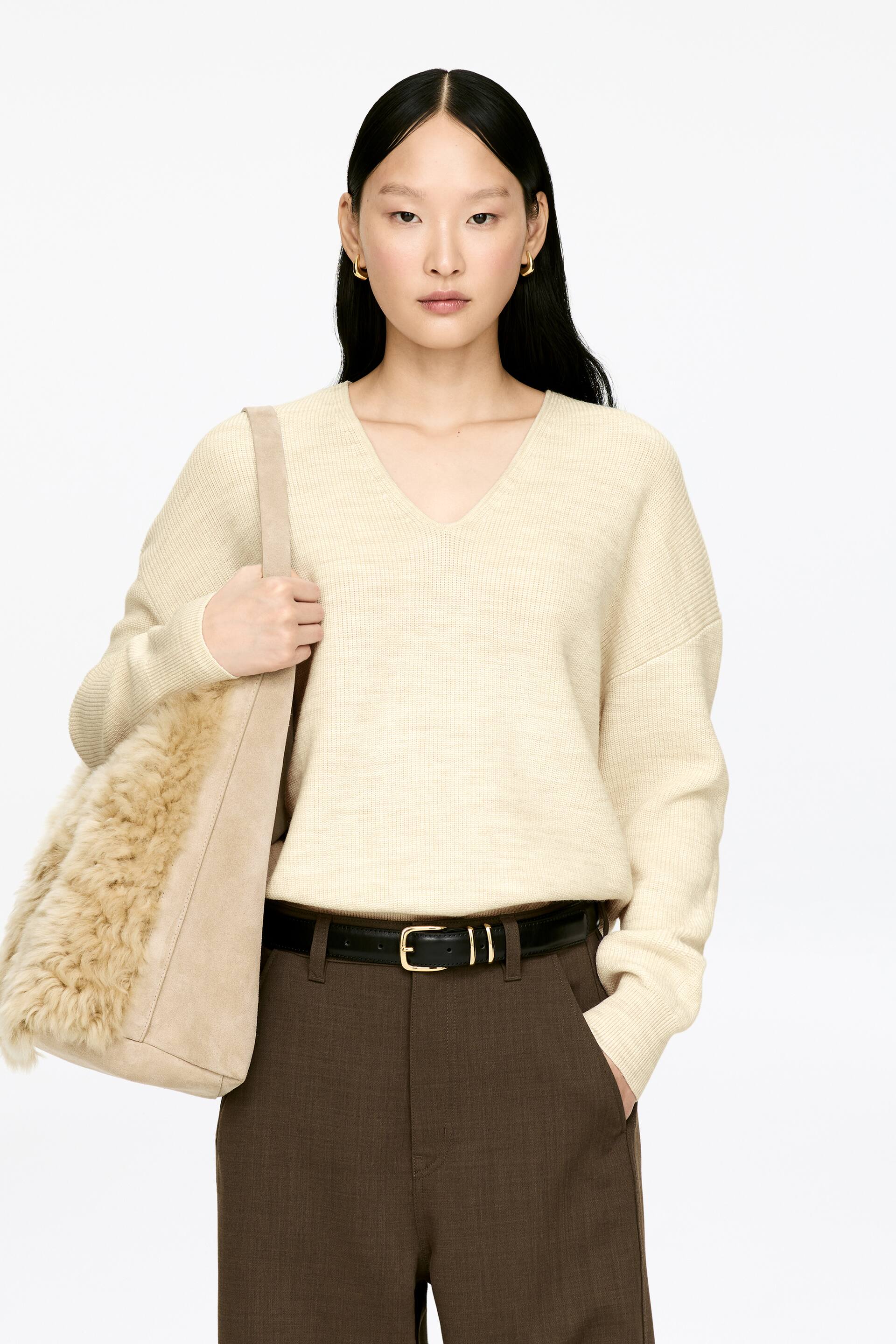 Zobacz większy obraz: Modelka ma na sobie kremowy sweter z dekoltem w serek, z prążkowanymi mankietami i dołem. Sweter jest stylizowany na włożony w brązowe spodnie z czarnym paskiem, a do swetra noszona jest beżowa torebka na ramię z imitacją futra.